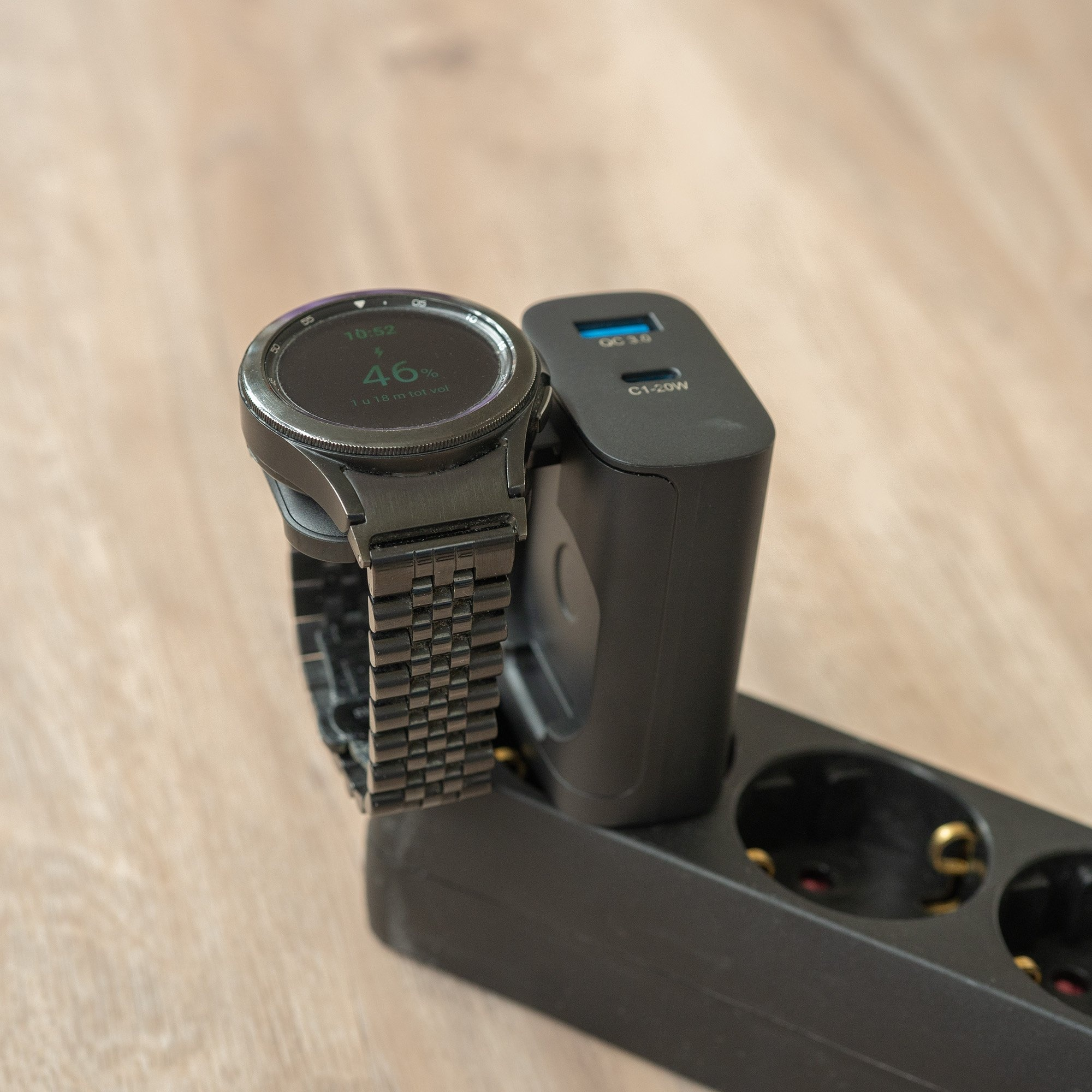 Eine schwarze Smartwatch mit 46% Akku lädt an einem Netzteil an einer Steckdosenleiste.