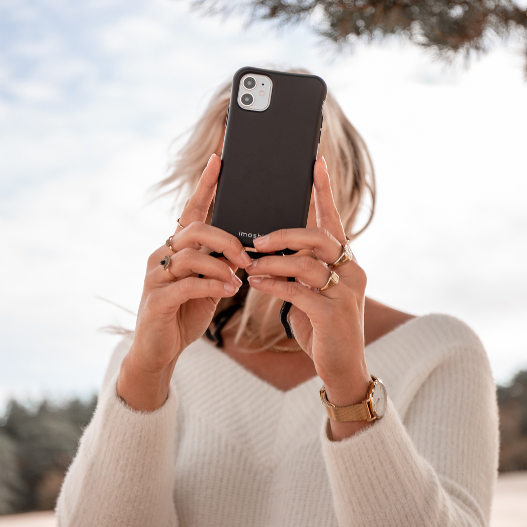 Vrouw met blond haar houdt zwarte telefoon voor gezicht, draagt witte trui.
