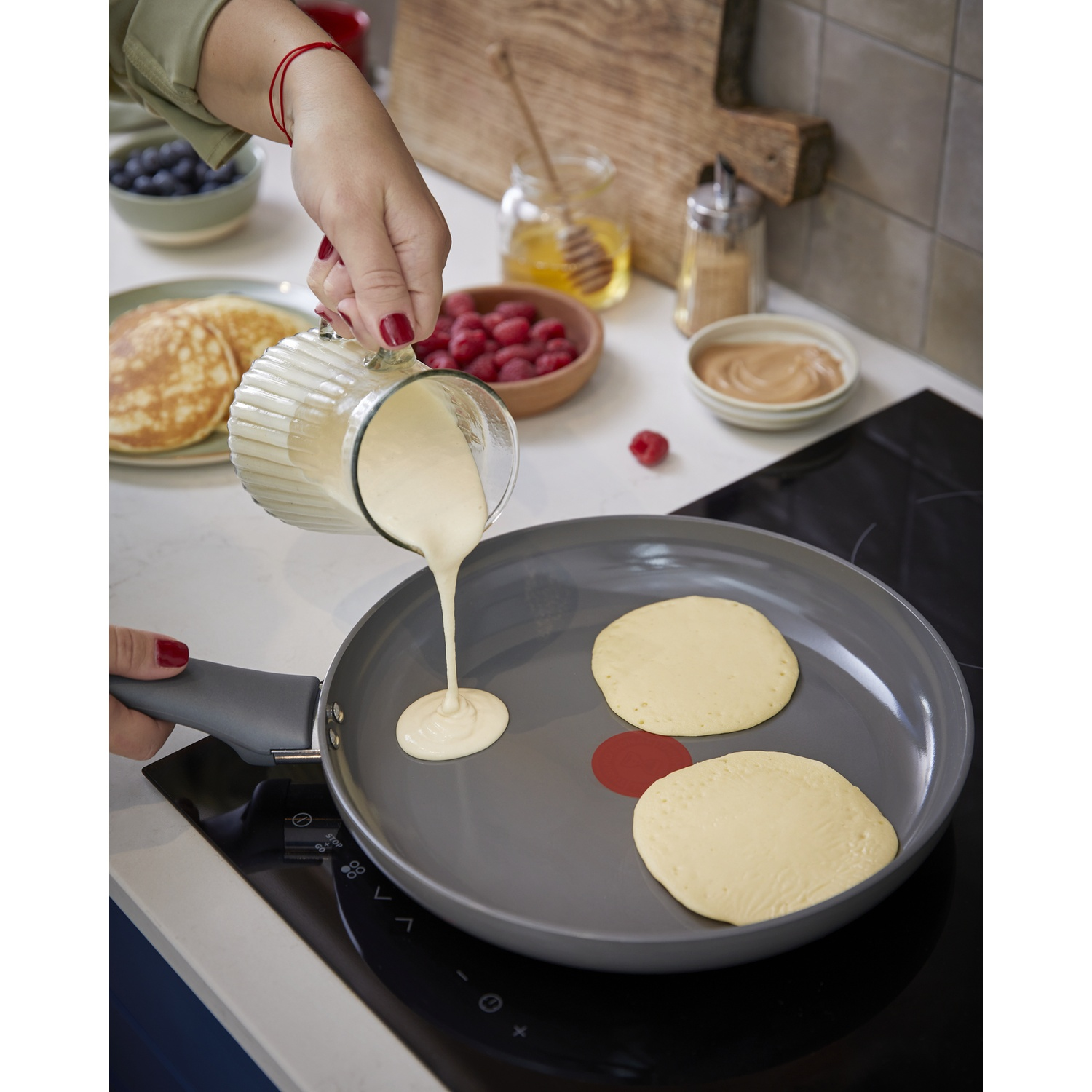 Una mano vierte masa en una sartén para cocinar panqueques. Hay bayas al fondo.