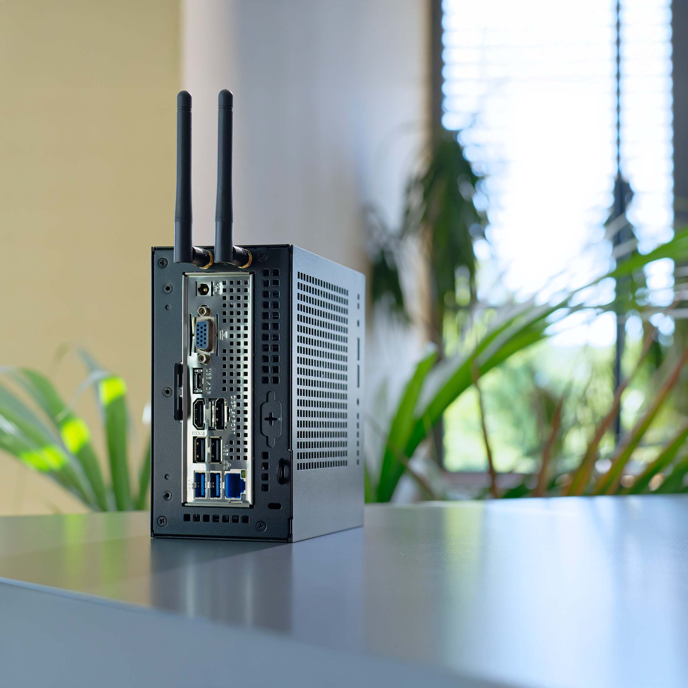 Schwarzer Mini-PC mit Antennen, Anschlüsse sichtbar. Grüne Pflanzen u. Jalousien im Hintergrund.