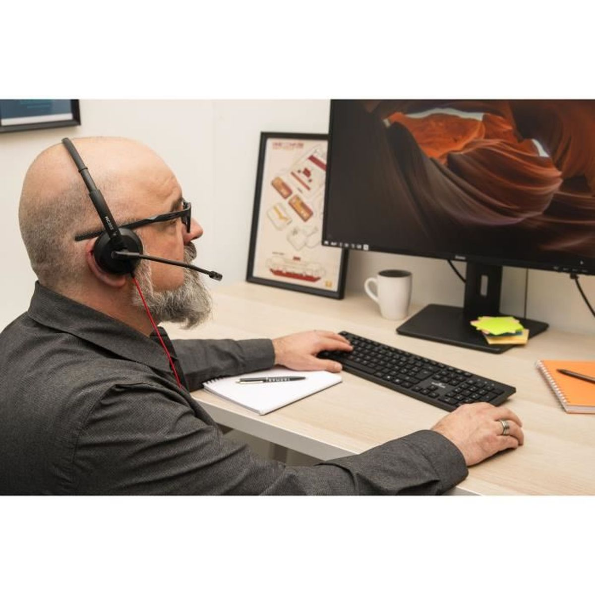 Mann mit Headset am Schreibtisch, der eine Tastatur benutzt. Ein Monitor zeigt eine Schlucht.