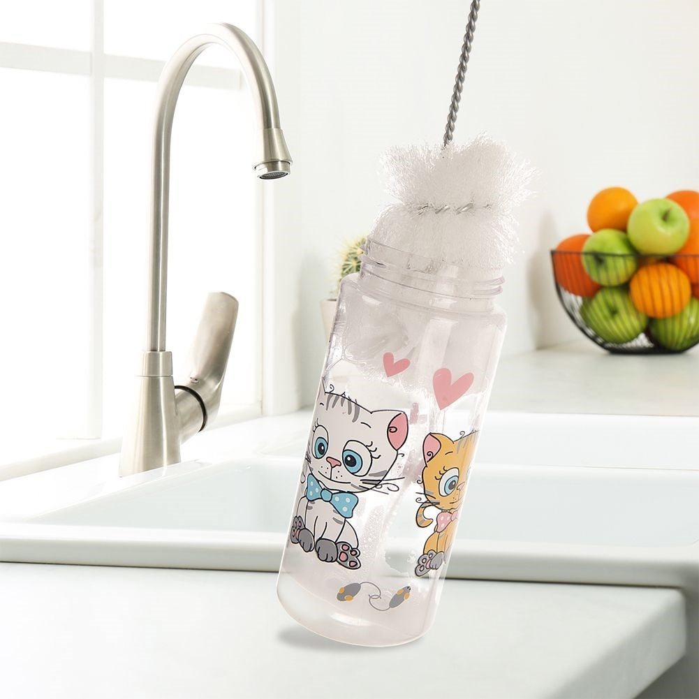 Un biberón con diseños de gatos se limpia con un cepillo, al lado de fruta en un bol.