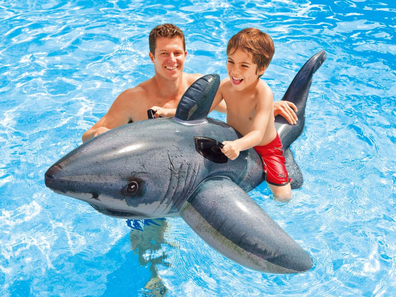 Hombre y niño montan un tiburón inflable en piscina.