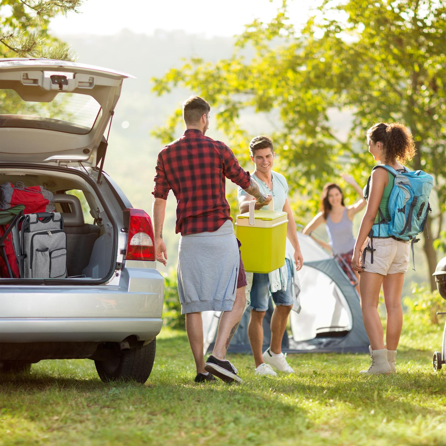 Gente descargando equipo de camping de un coche, con tienda y parrilla al fondo.