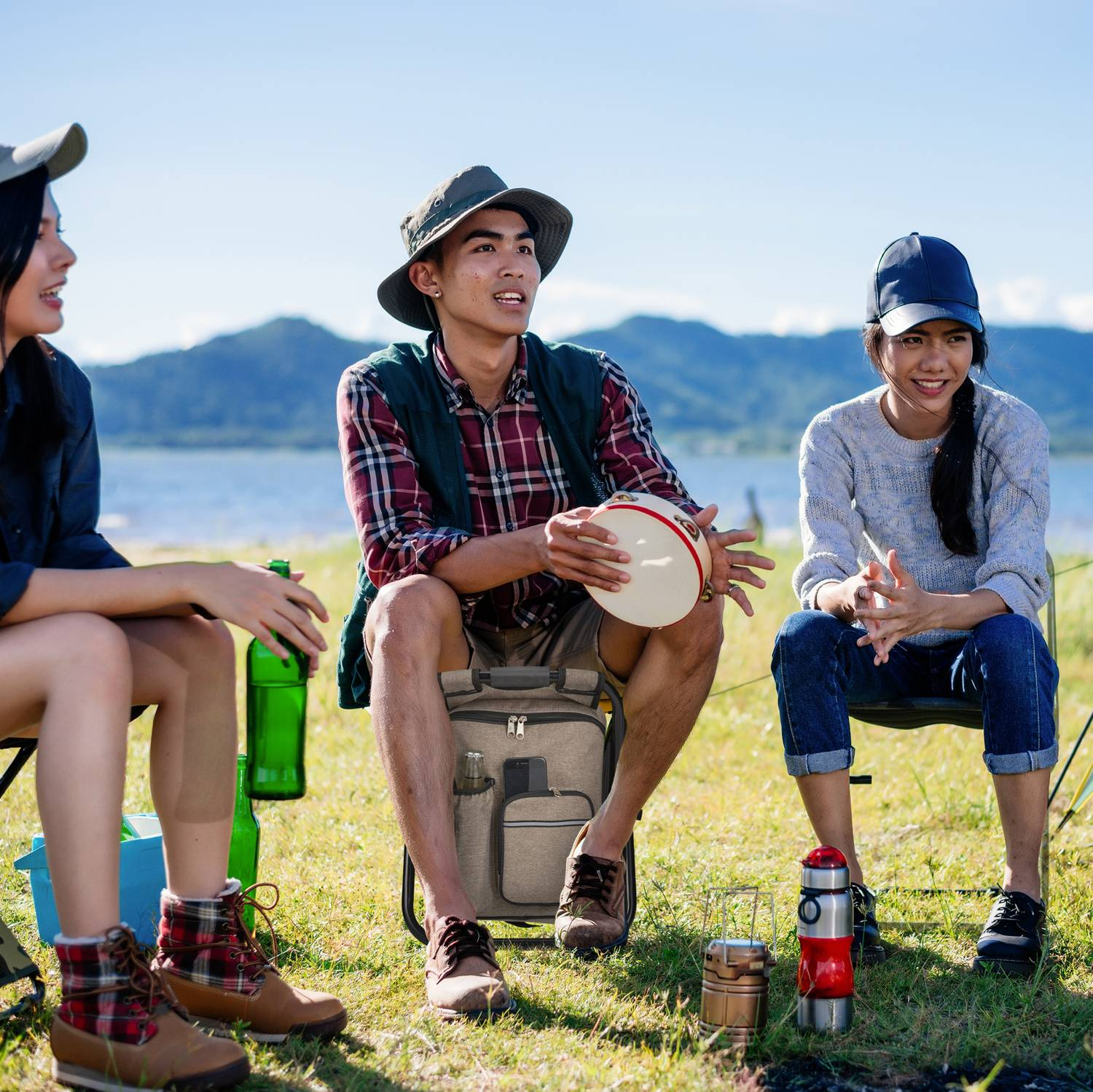 Tres amigos disfrutando un picnic junto a un lago. Uno sostiene una pandereta. Montañas al fondo.