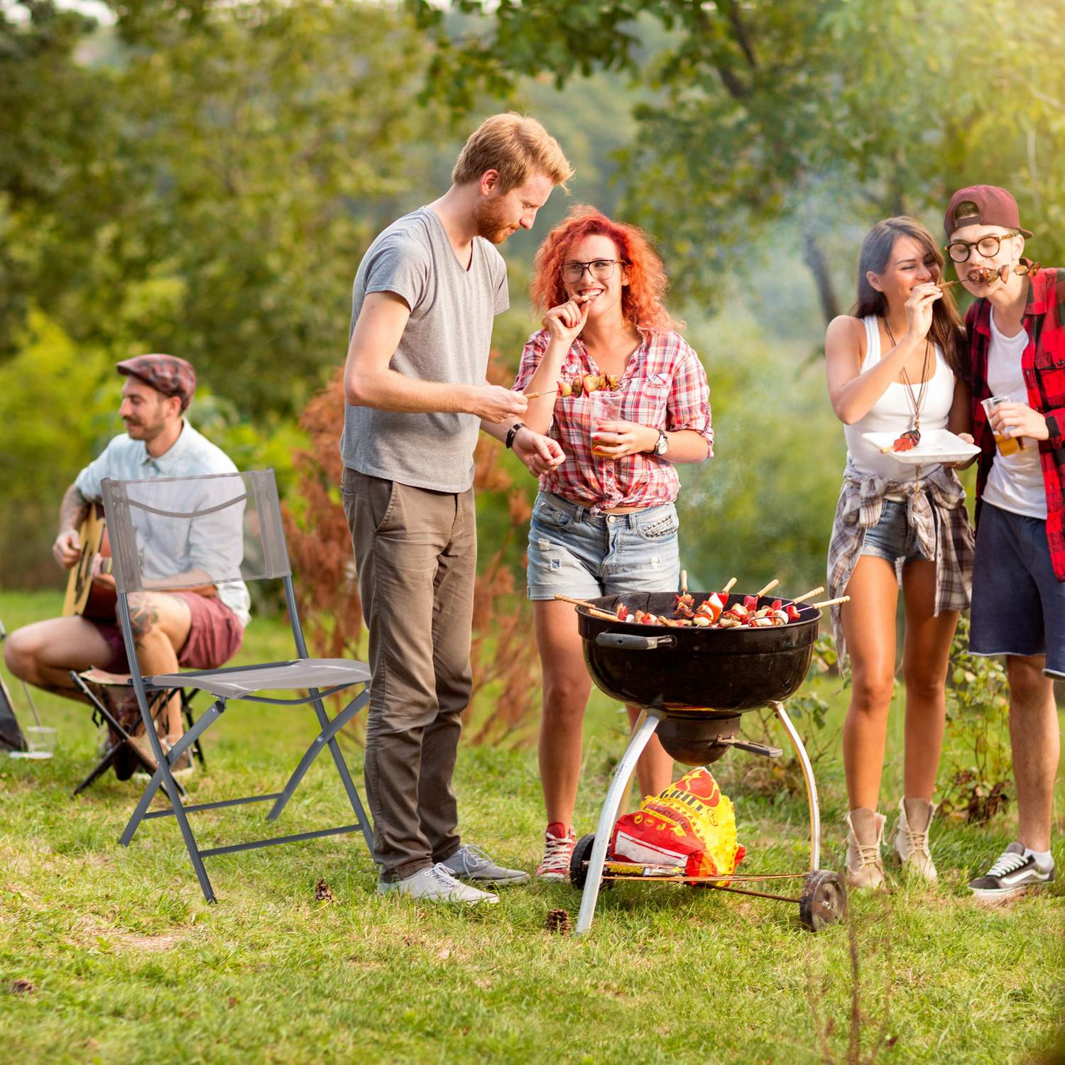 Amigos hacen barbacoa al aire libre, uno toca la guitarra mientras otros comen brochetas.