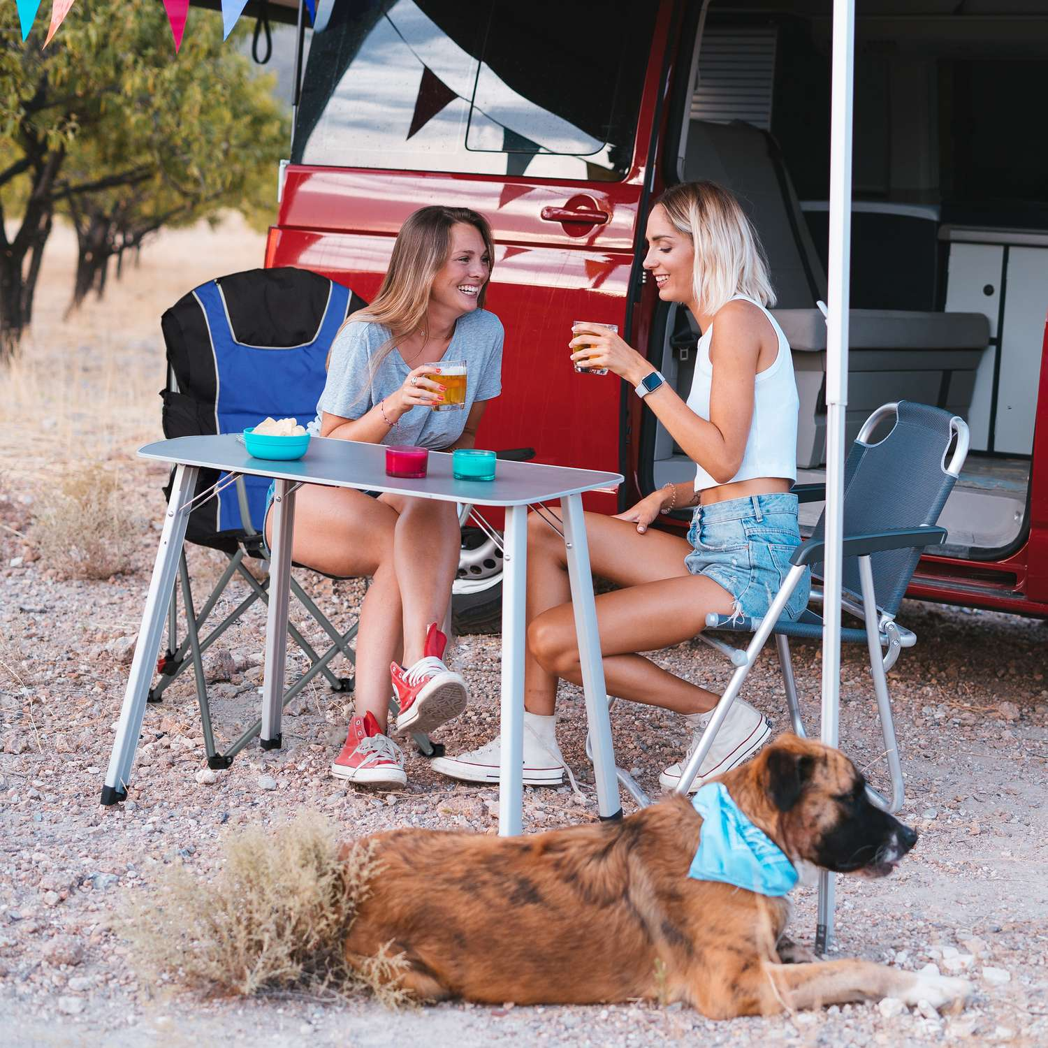 Dos mujeres beben afuera, un perro yace cerca. Una autocaravana está al fondo.