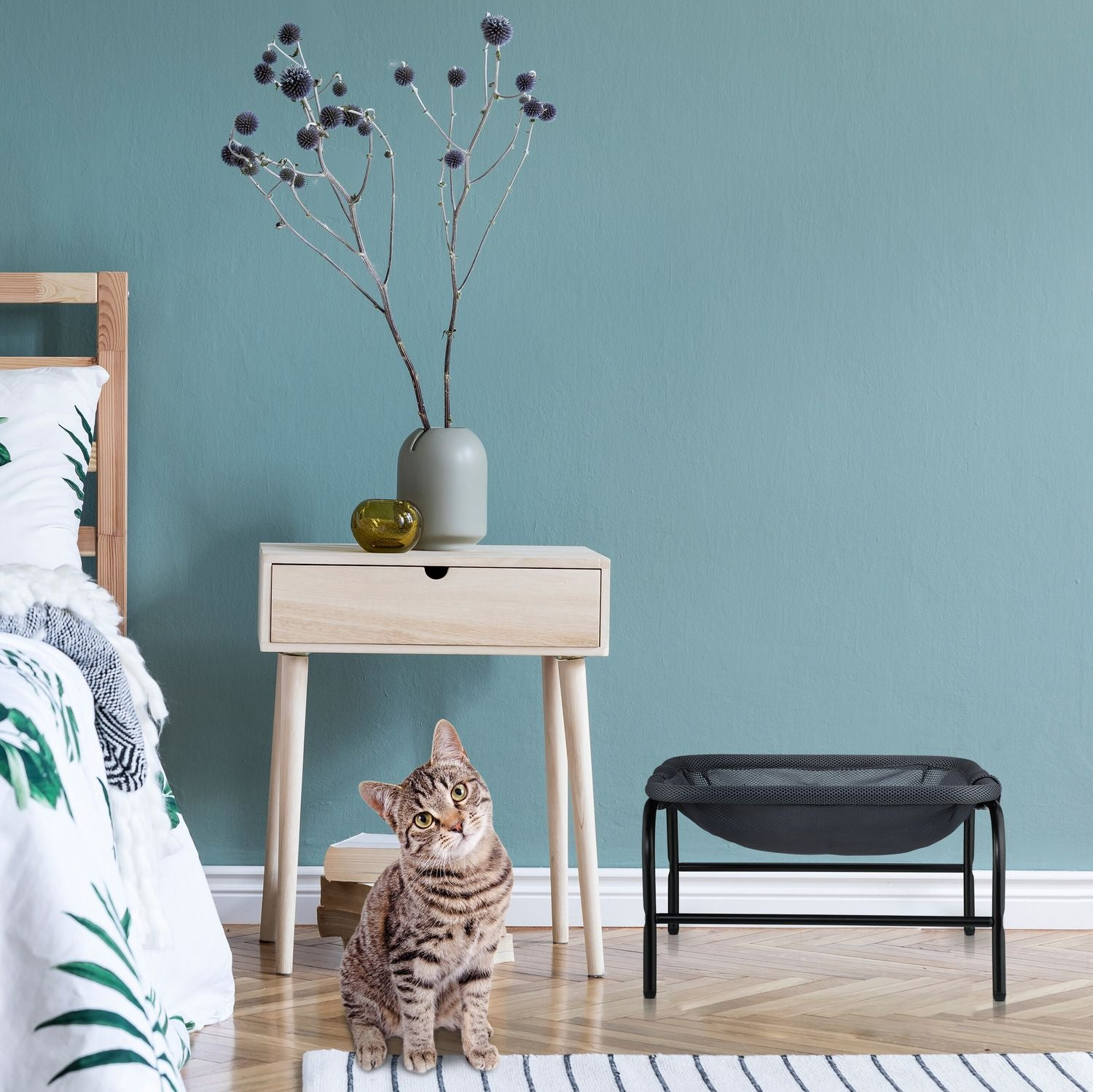 Un gato se sienta junto a una cama con ropa de cama floral. Se ven una mesita de noche y una cama para gatos.