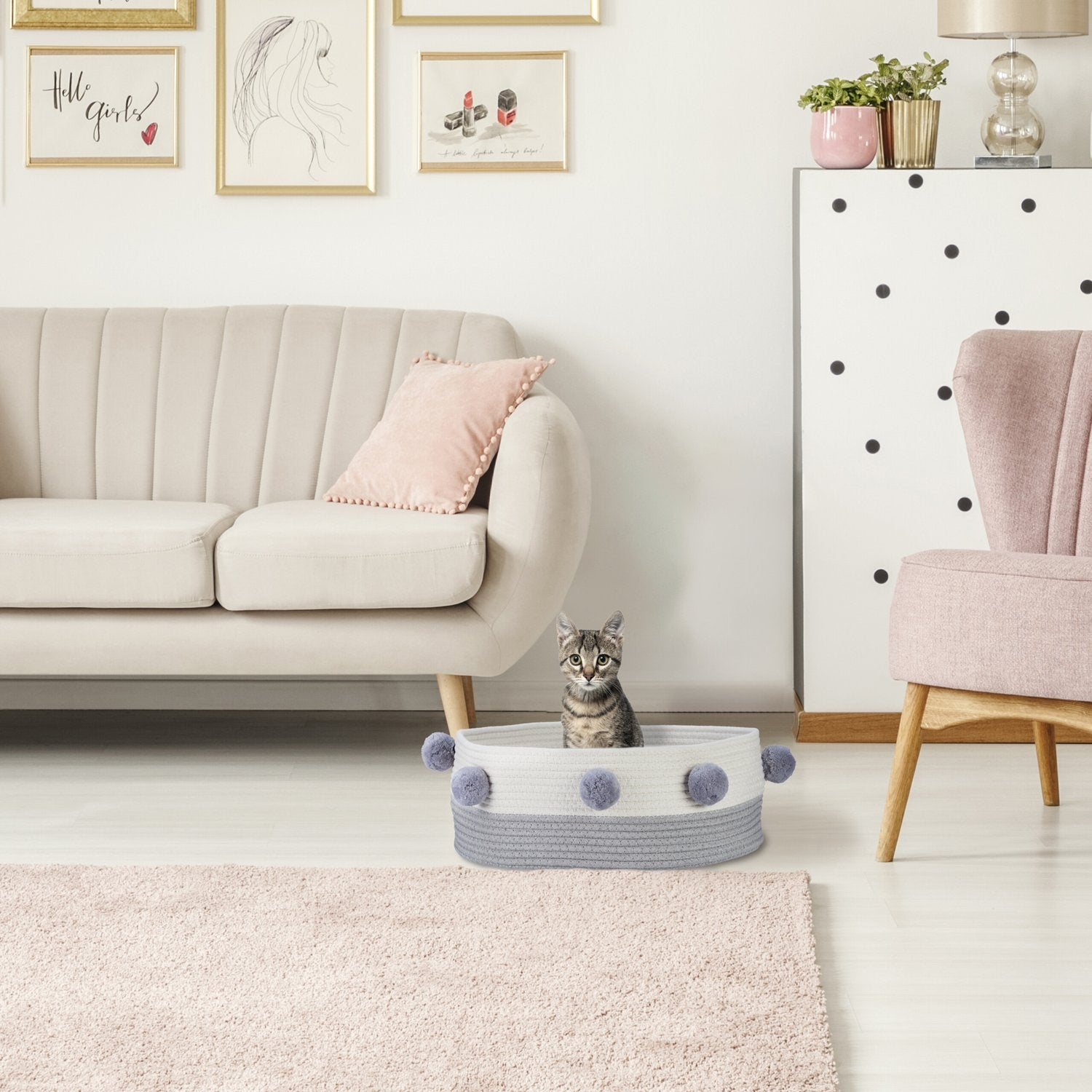 Un gato se sienta en una cesta con pompones grises junto a un sillón rosa.