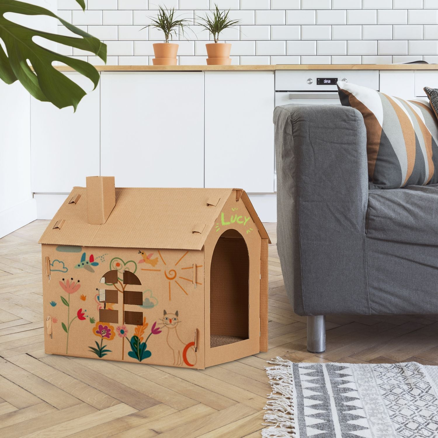 Una casa de cartón para gatos con dibujos coloridos está sobre un suelo de madera junto a un sofá gris.