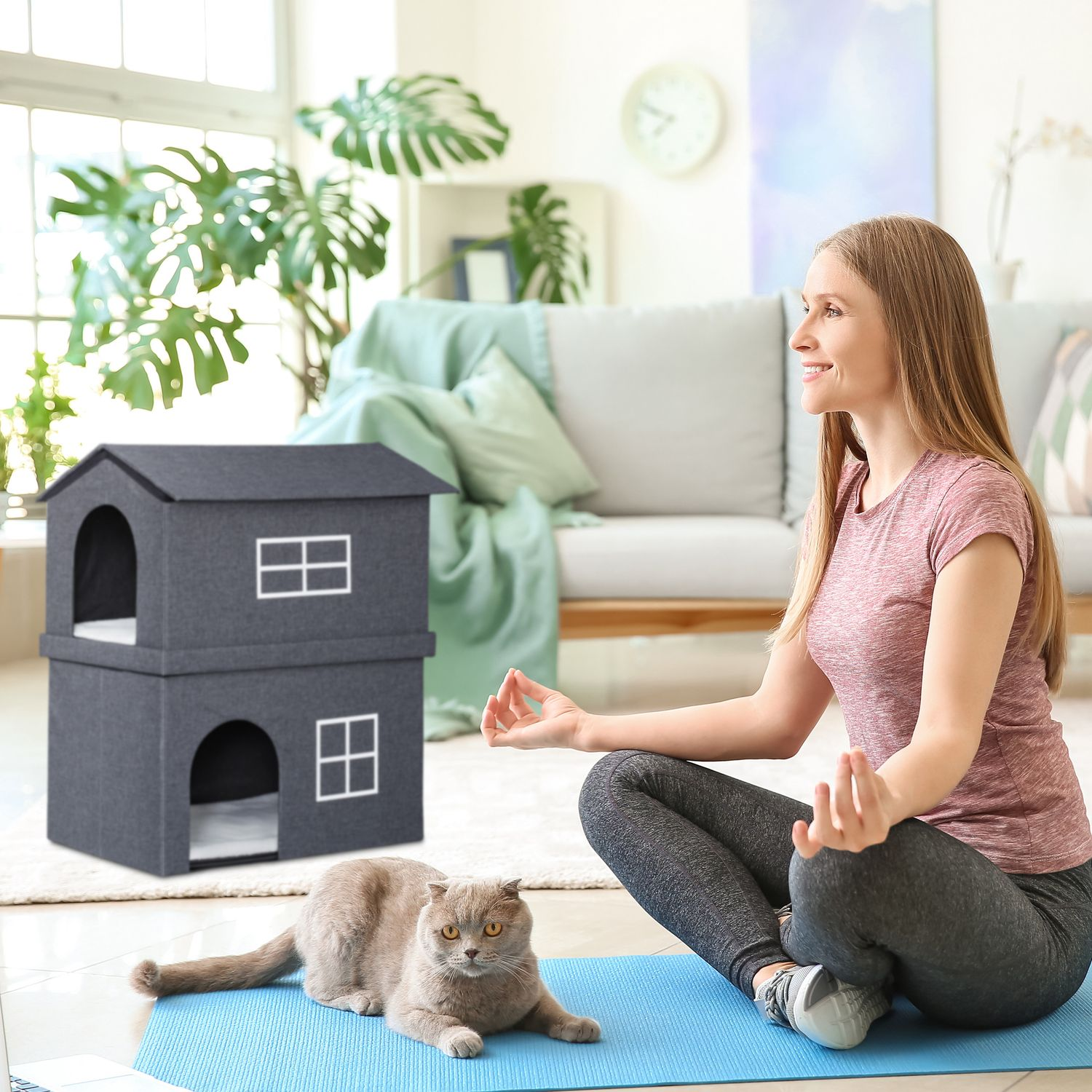Mujer meditando junto a una casa para gatos gris y un gato.