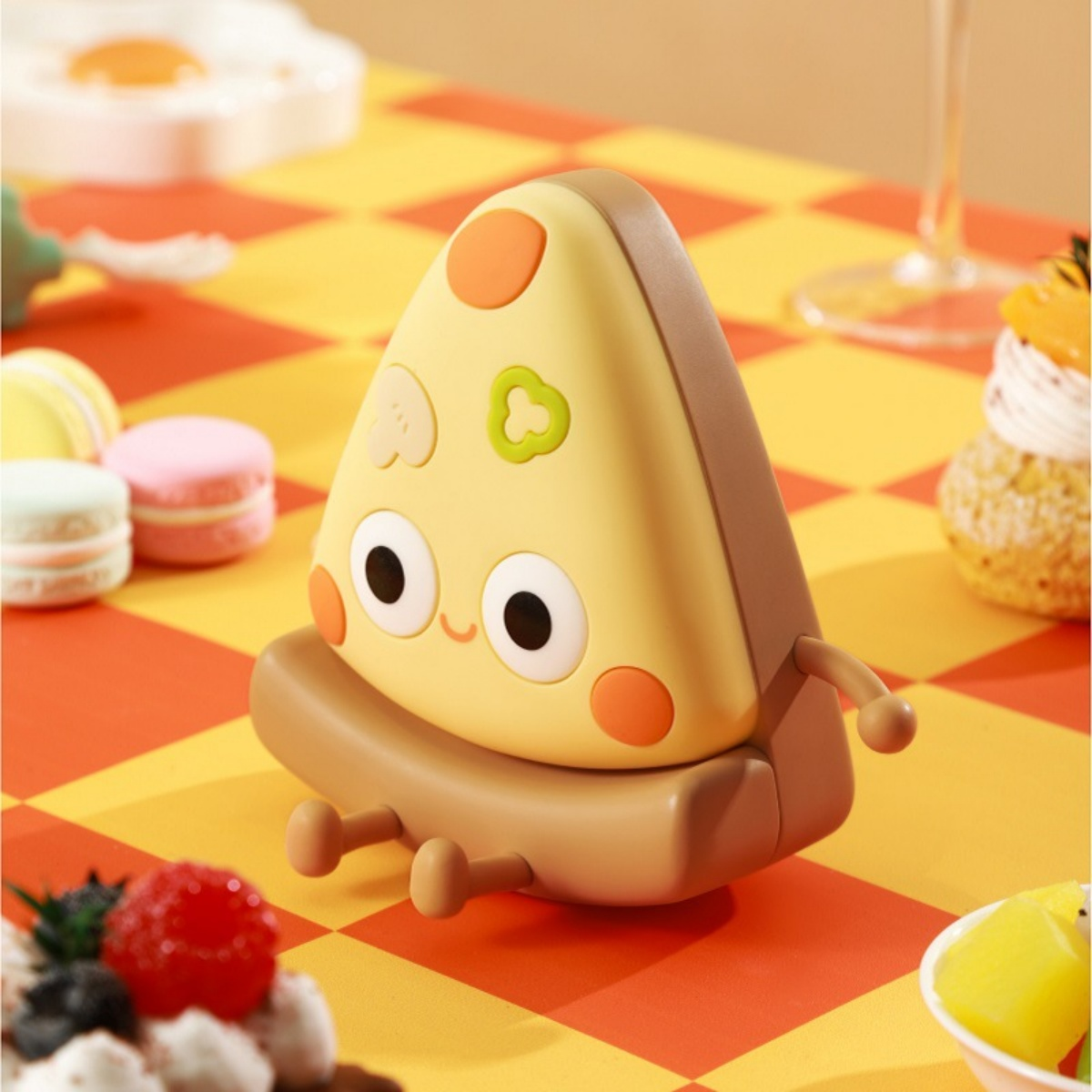 Eine lustige Pizzastück-Figur sitzt auf einem karierten Tisch mit Macarons und Desserts.