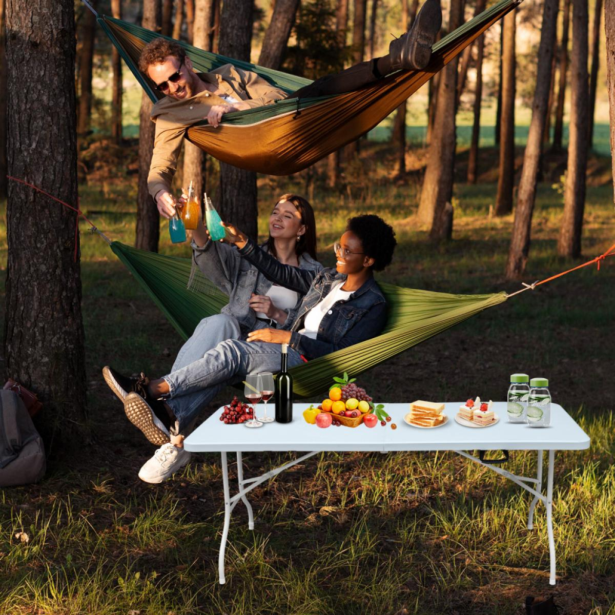 Freunde stoßen in Hängematten an; Picknicktisch mit Obst, Sandwiches, Kuchen.