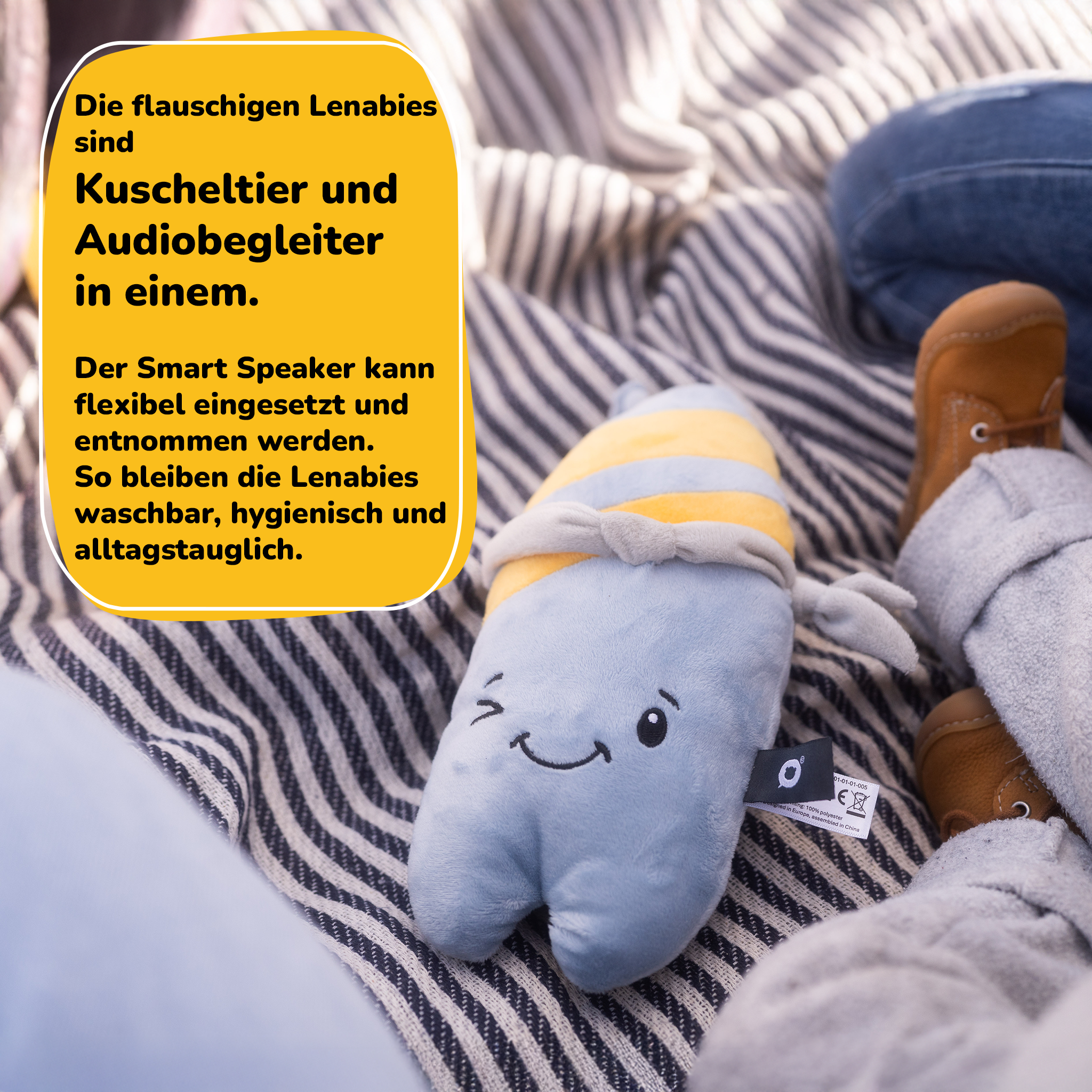 Ein hellblaues Plüschtier mit gelben Streifen und einem aufgenähten Gesicht liegt auf einer gestreiften Decke.