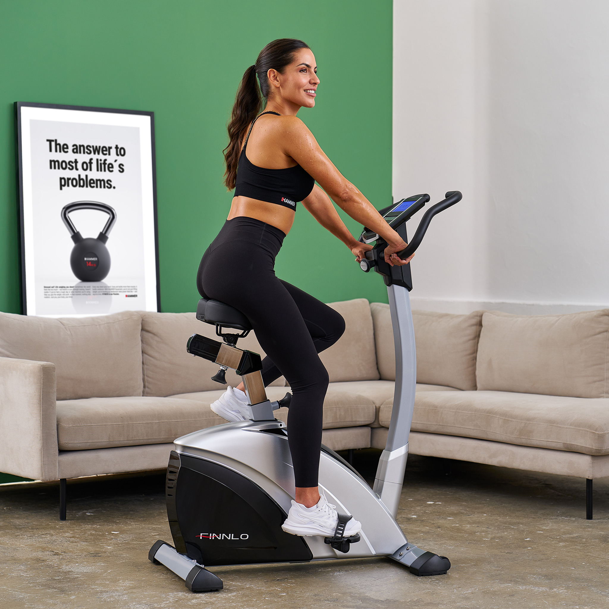 Frau trainiert auf einem Heimtrainer vor einem Sofa und einem Bild mit Kettlebell.