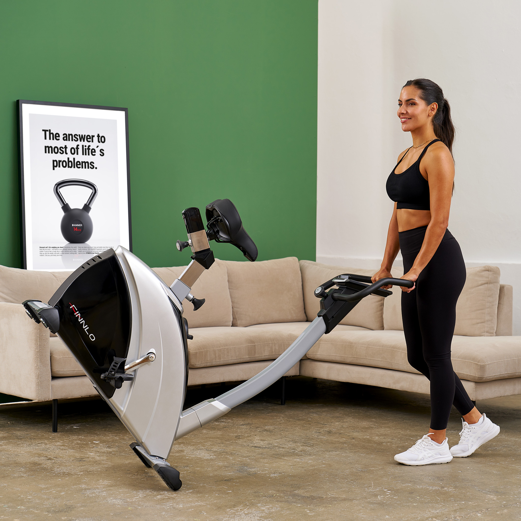 Eine Frau in Sportkleidung steht neben einem silbernen Heimtrainer. Ein Poster hängt an der Wand.