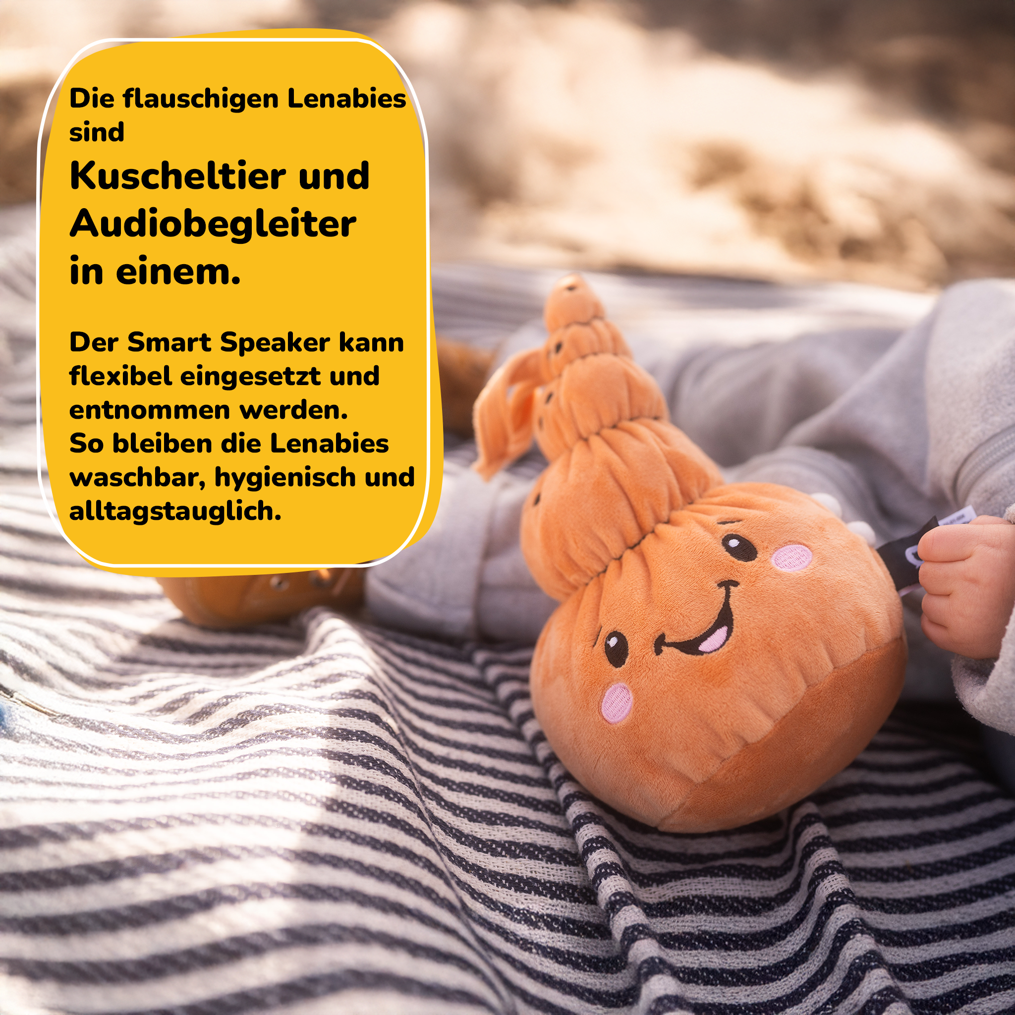 Ein orangefarbenes Plüschtier mit Gesicht, auf einer gestreiften Decke, gehalten von einer Babyhand.