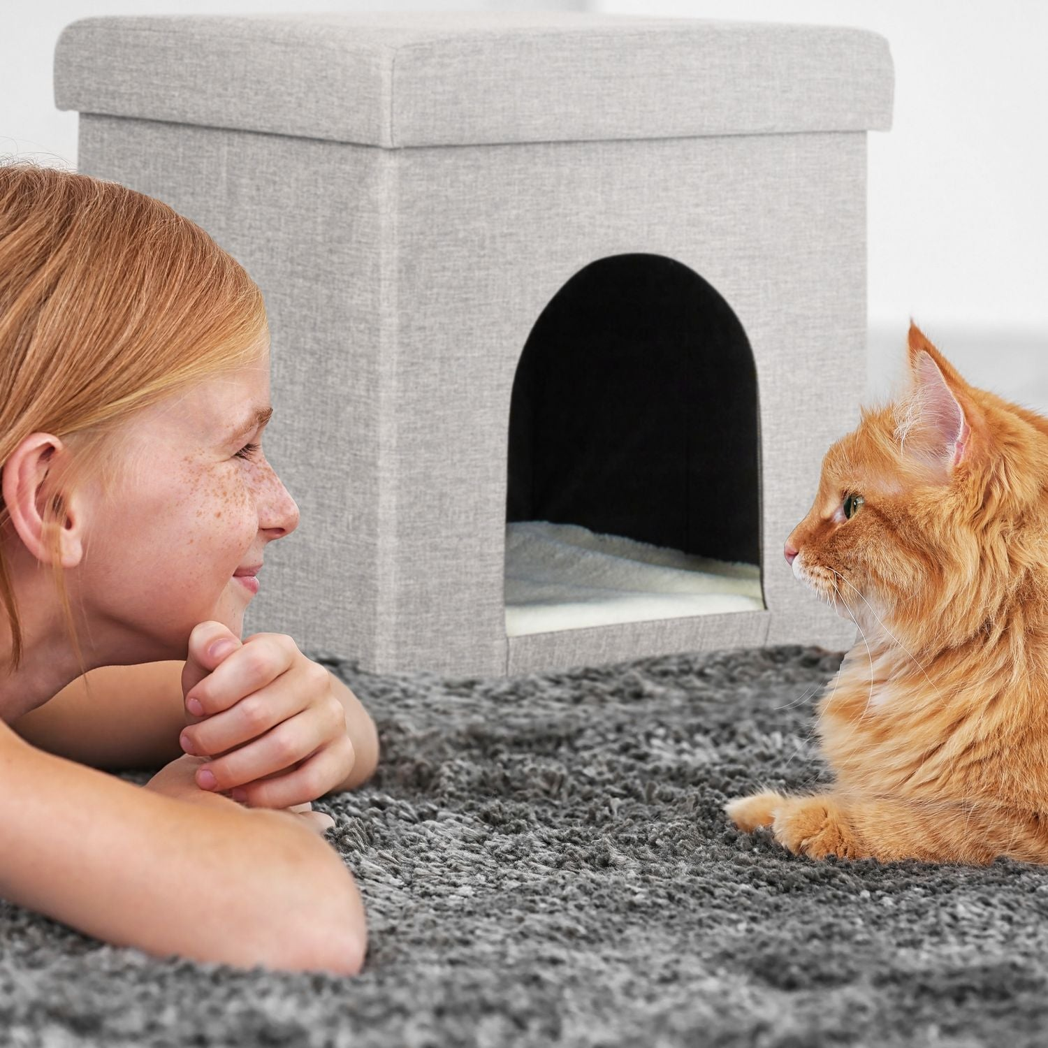 Una niña y un gato naranja se miran cerca de una casa para gatos gris.