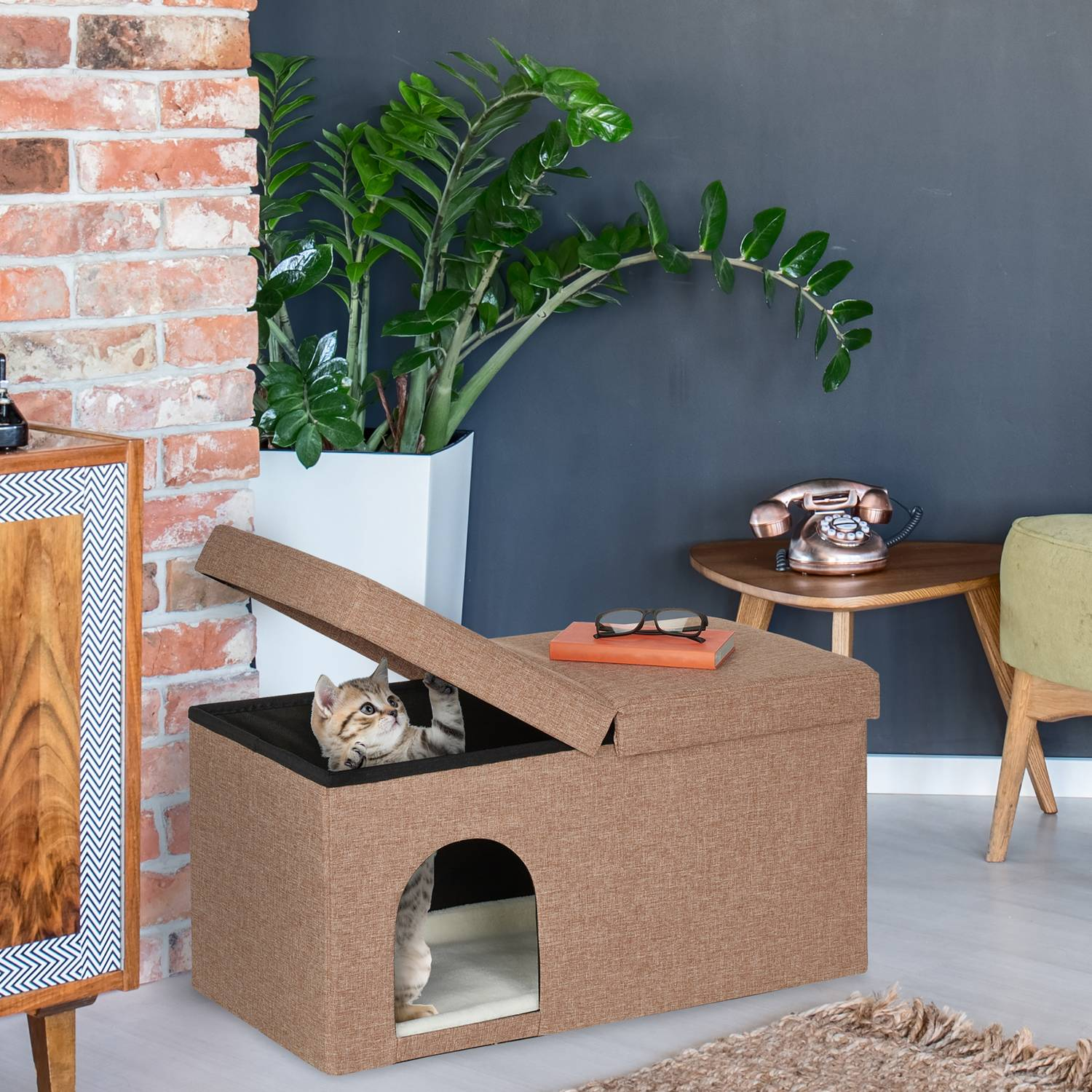 Un gato se sienta dentro de una casa para gatos marrón junto a una pared de ladrillos y una planta.