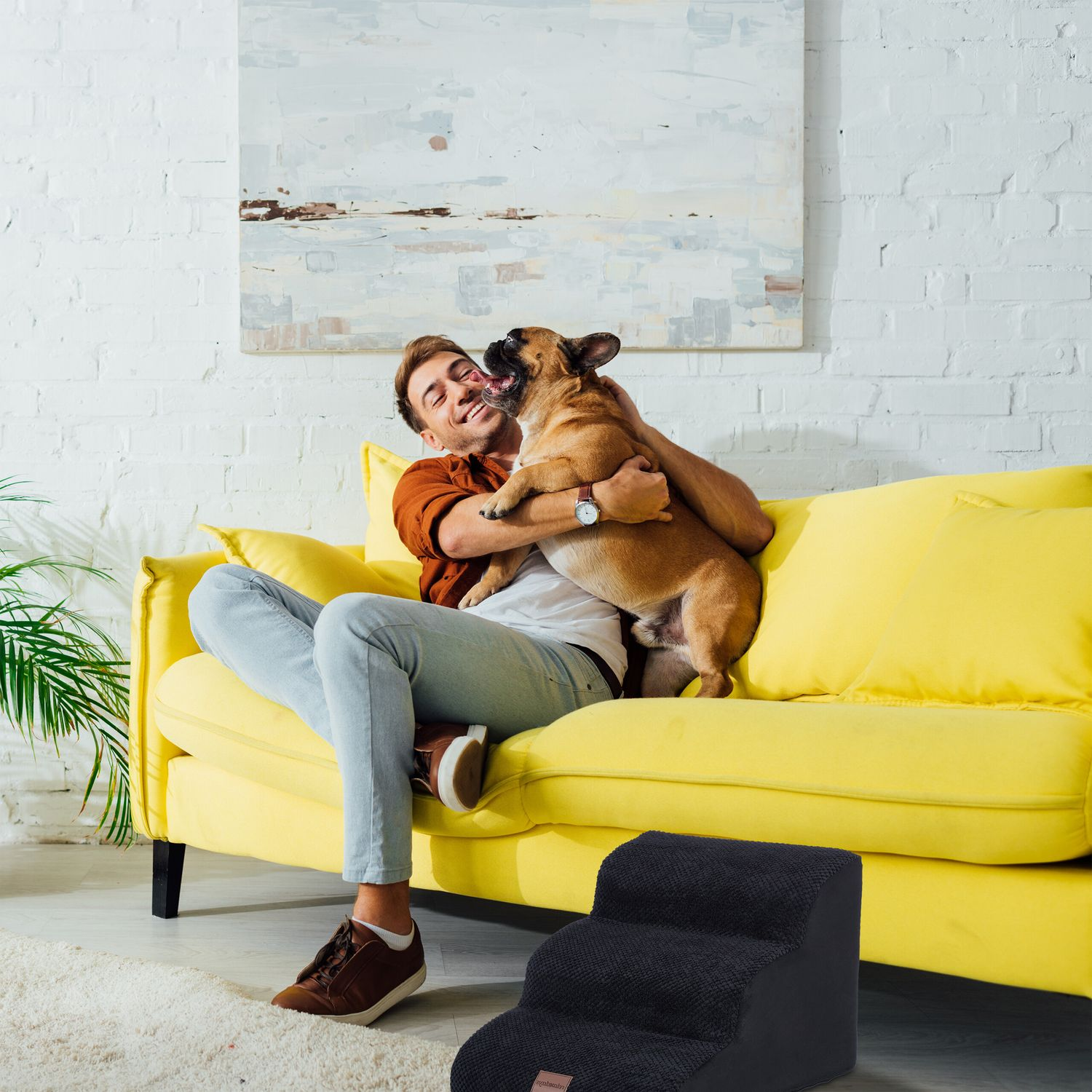 Un hombre abraza a su bulldog francés en un sofá amarillo.