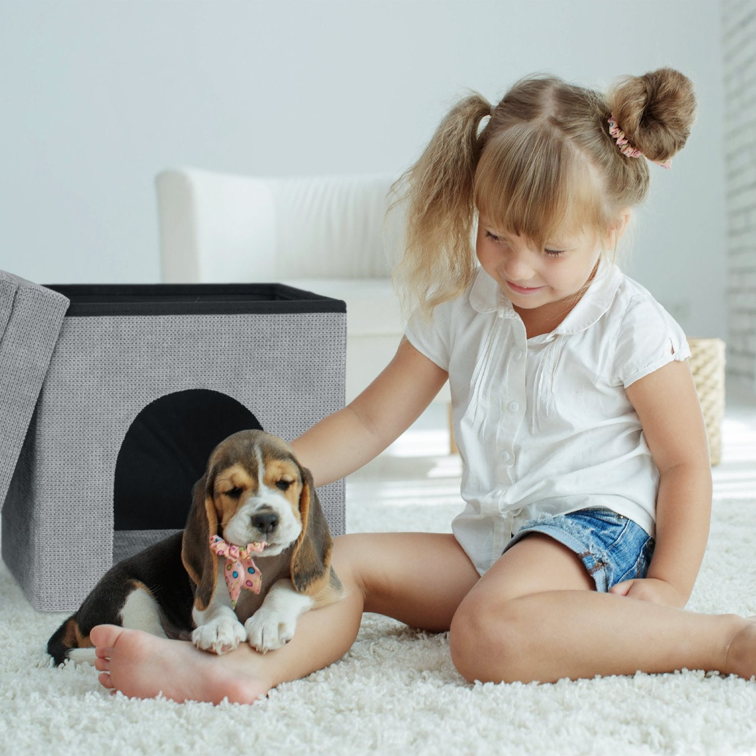 Un cachorro beagle con lazo se sienta en el regazo de una niña junto a una casa para mascotas gris.