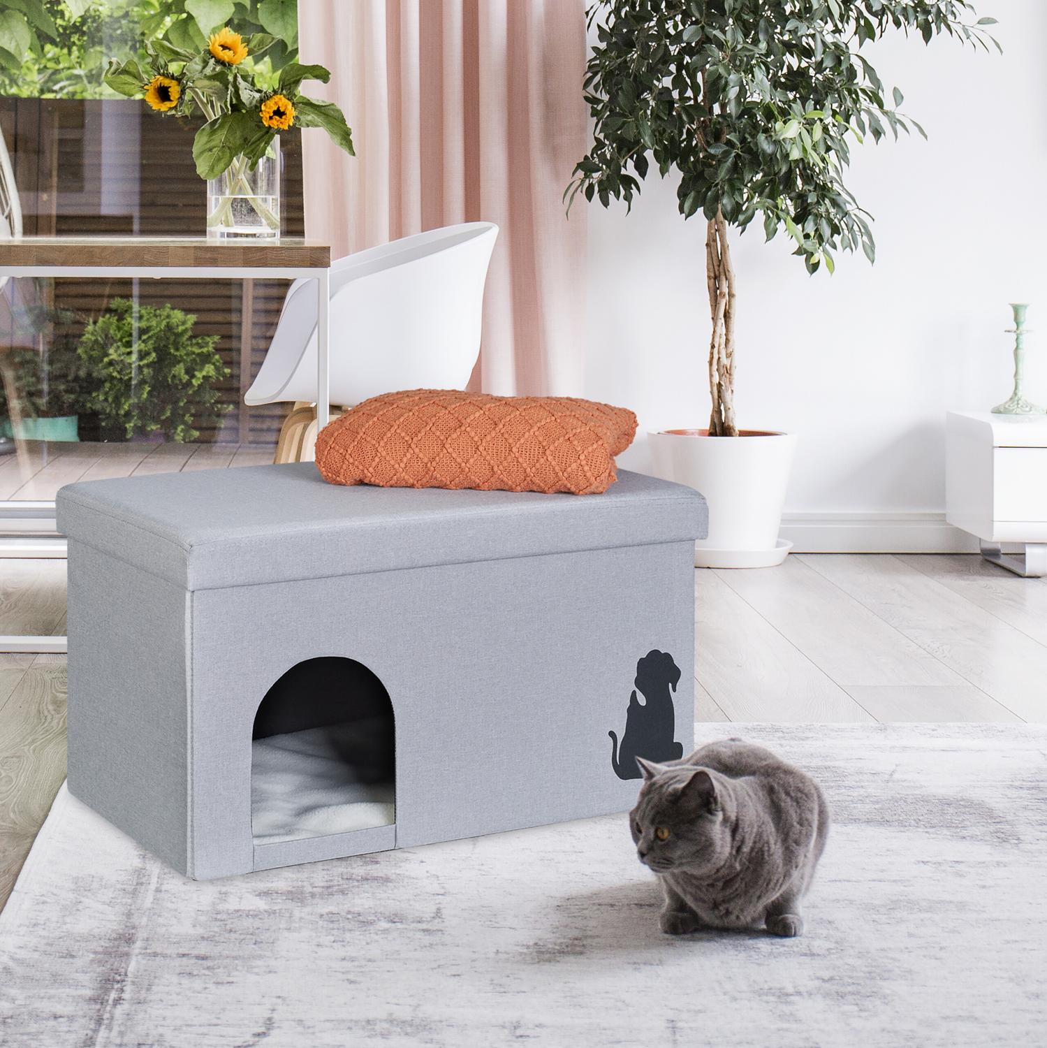 Un gato gris se sienta en una alfombra cerca de una casa para mascotas gris con un cojín naranja encima.
