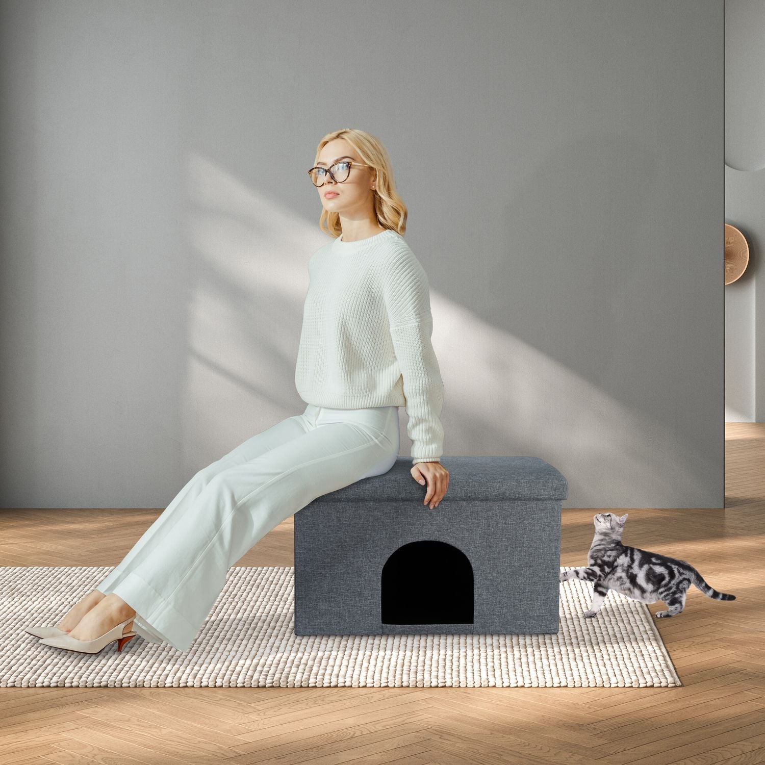 Una mujer se sienta en una casa para gatos gris, un gato juega a su lado.