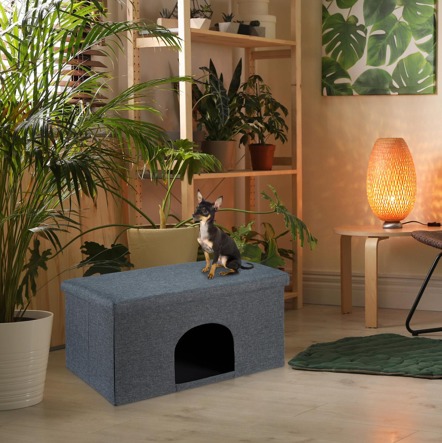Un perro pequeño se sienta sobre una casa gris para mascotas en una sala con plantas.