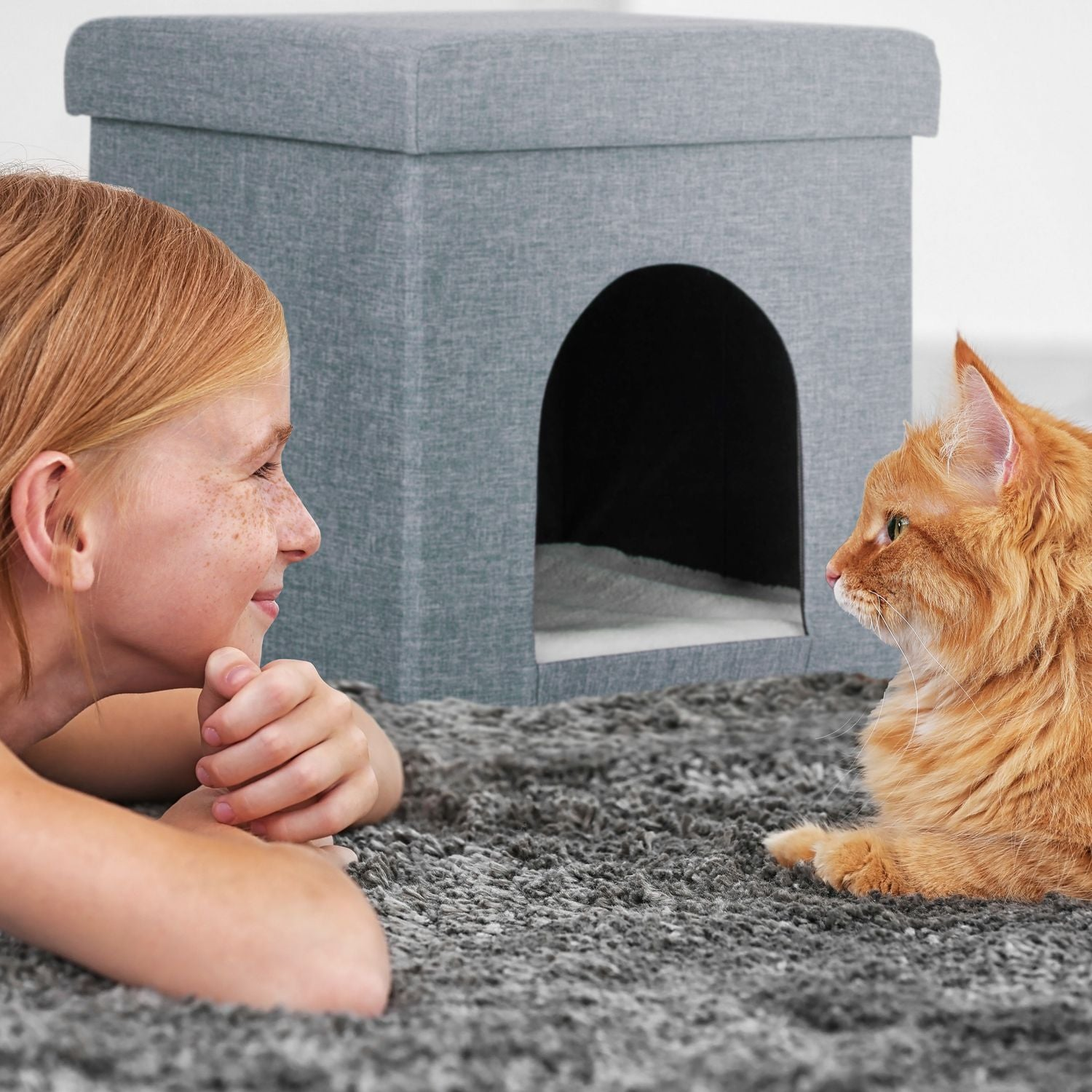 Una niña y un gato naranja se miran junto a una casa para gatos gris.