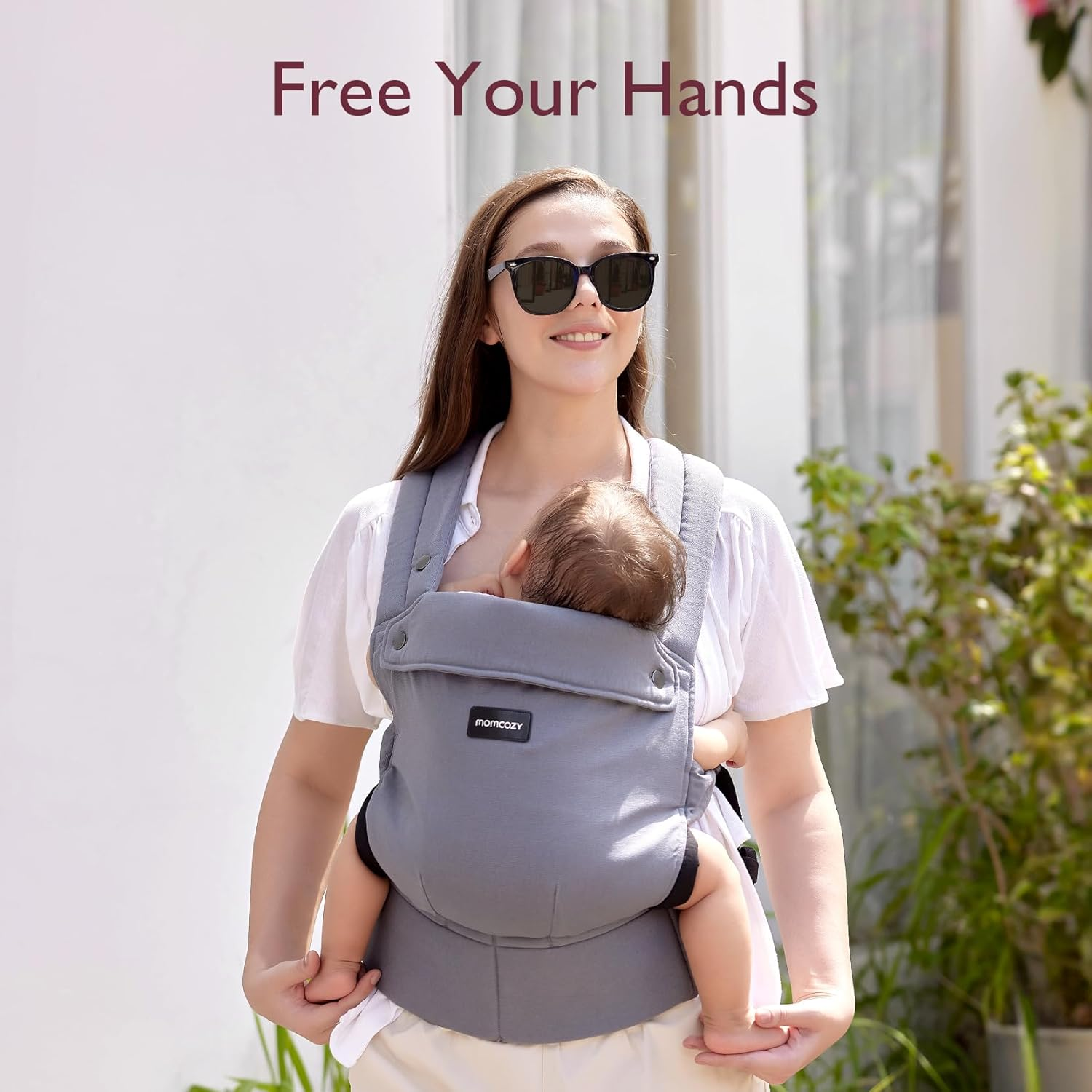 Una donna con occhiali da sole porta un bambino in un marsupio grigio.