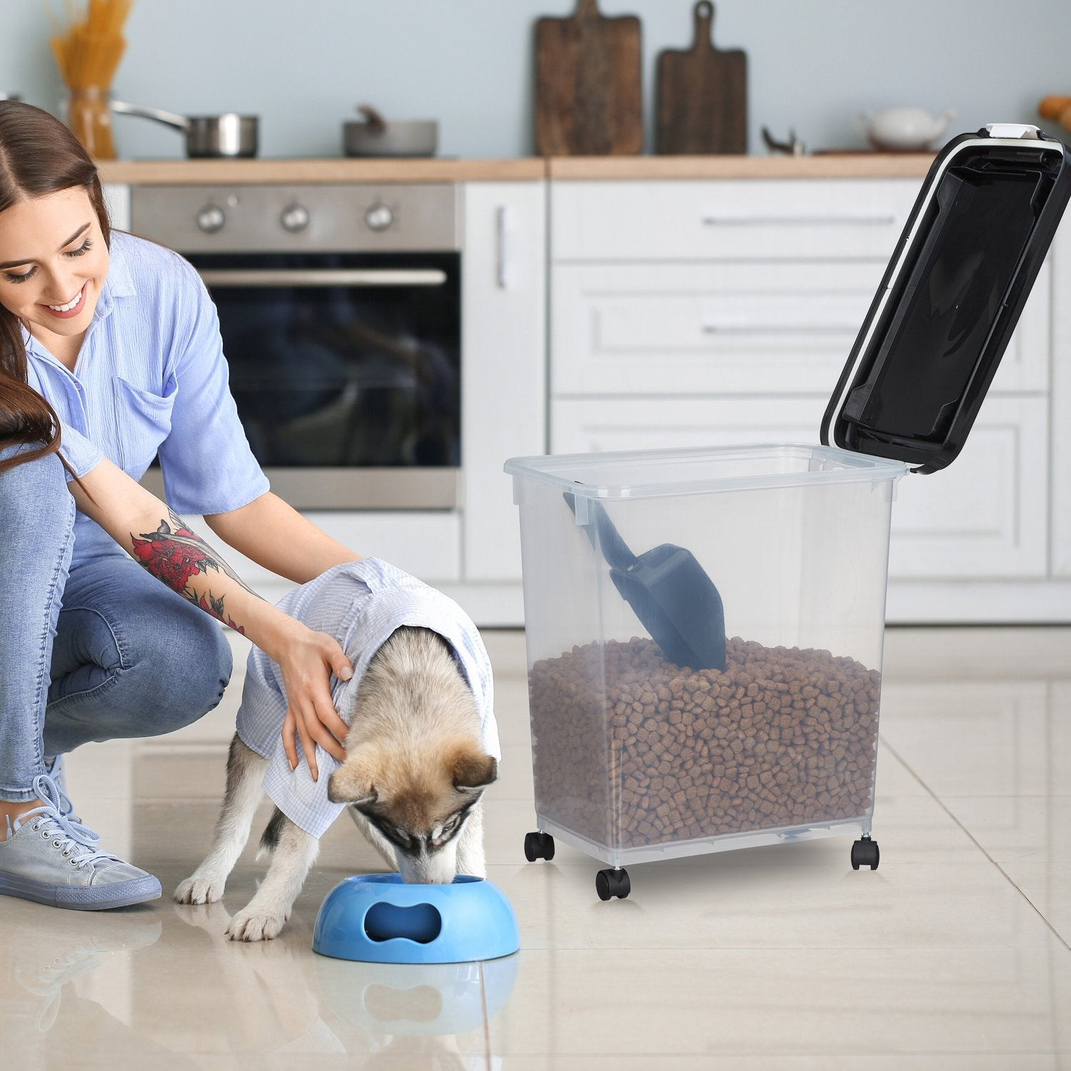 Mujer con camisa azul y jeans acaricia cachorro husky comiendo de cuenco azul junto a contenedor de comida.