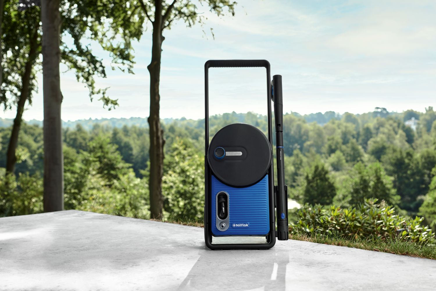 Blue pressure washer with black frame and hose reel sits on a patio before trees.