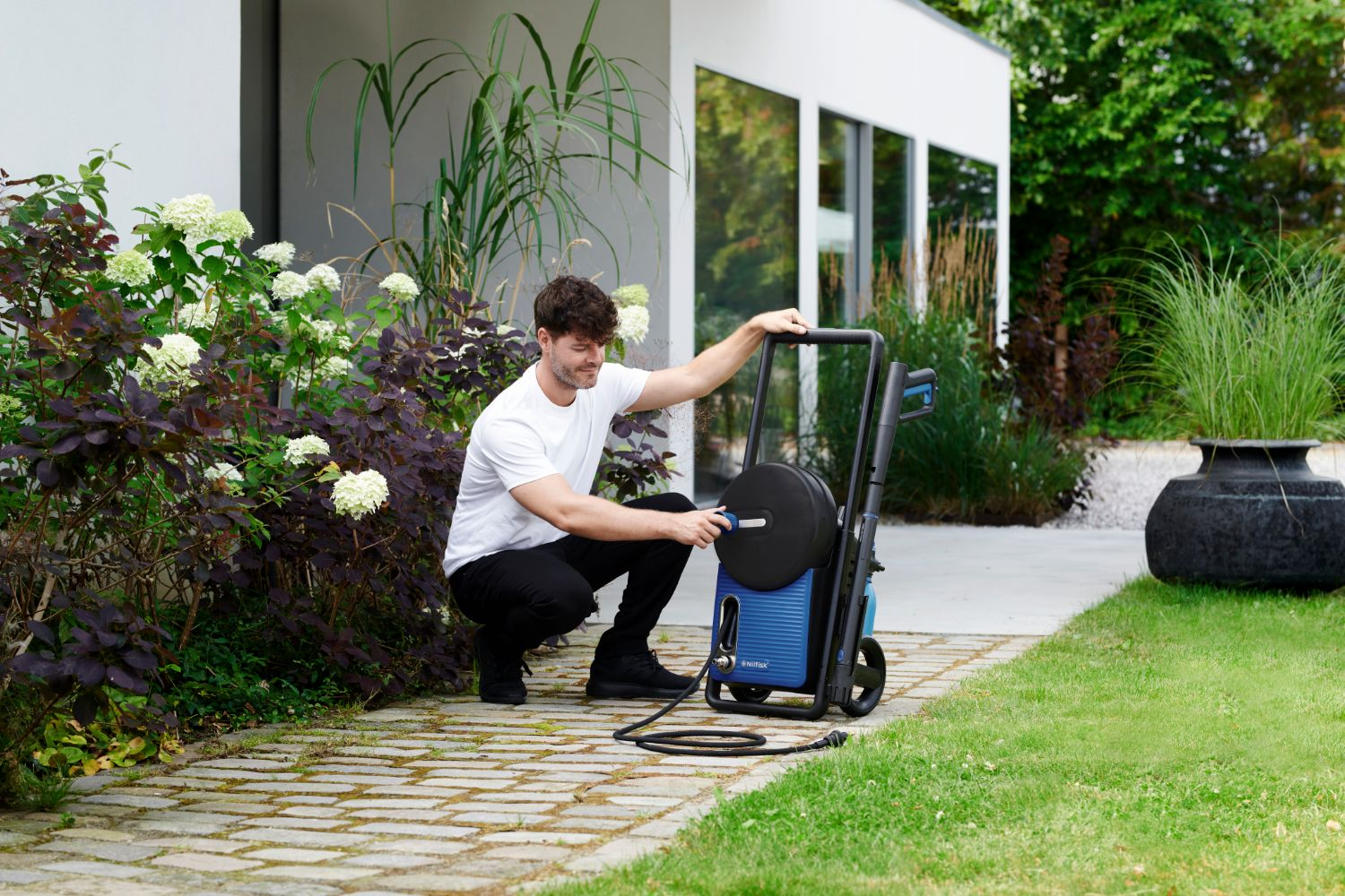 Man in white t-shirt crouching by blue pressure washer in a garden.