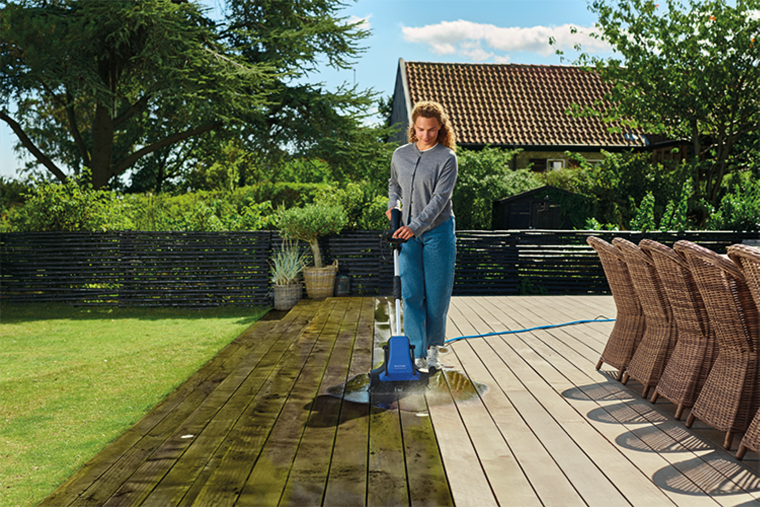Woman cleans wooden deck with pressure washer in garden.