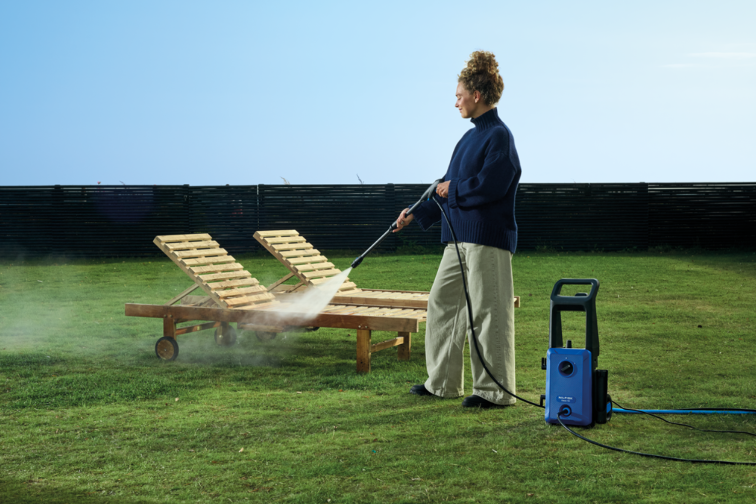 A woman cleans deck chairs with a pressure washer in a garden.