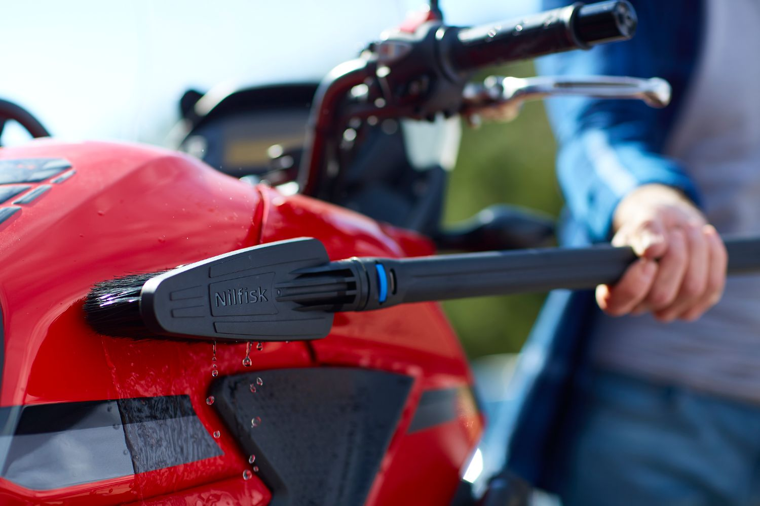 A hand holds a Nilfisk brush attachment cleaning a red motorcycle fairing.