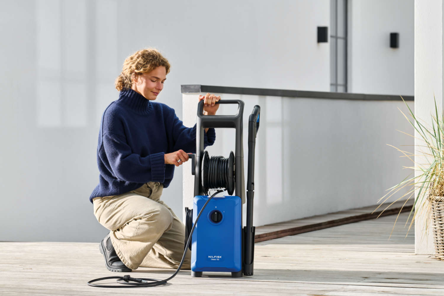Woman kneels beside a blue pressure washer with coiled hose.