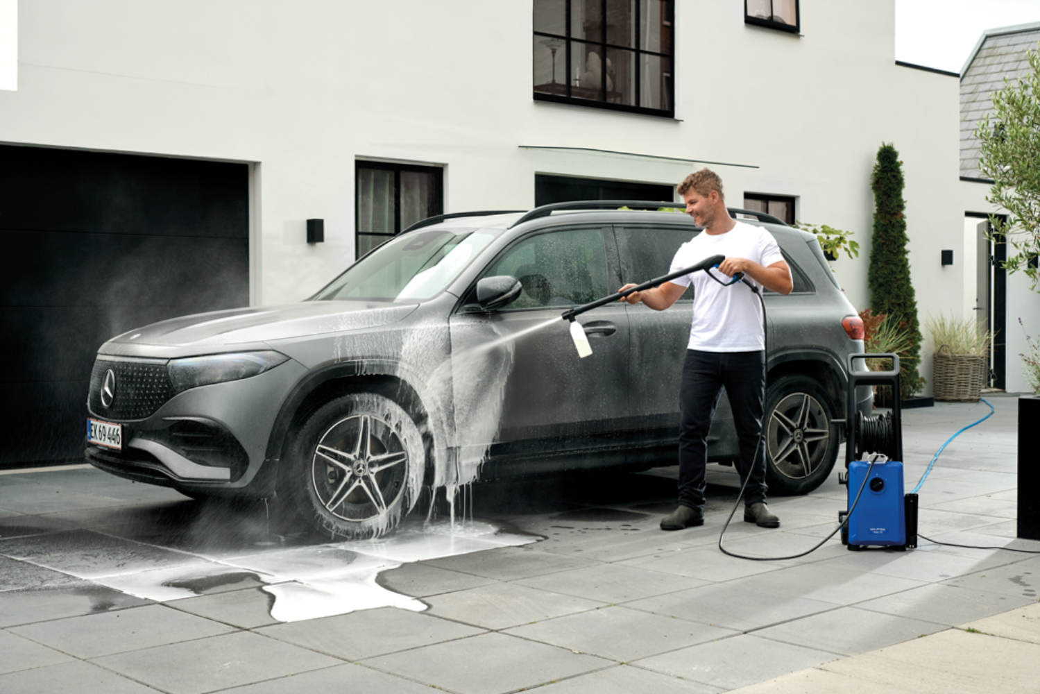 Man washes grey car with pressure washer in front of modern house.