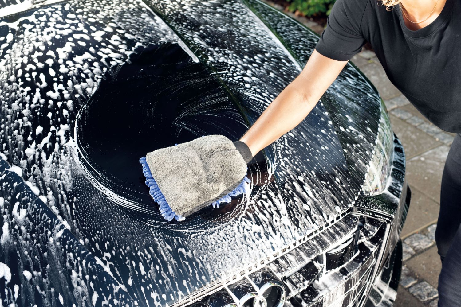 A hand wearing a wash mitt cleans the wet hood of a black car with suds.