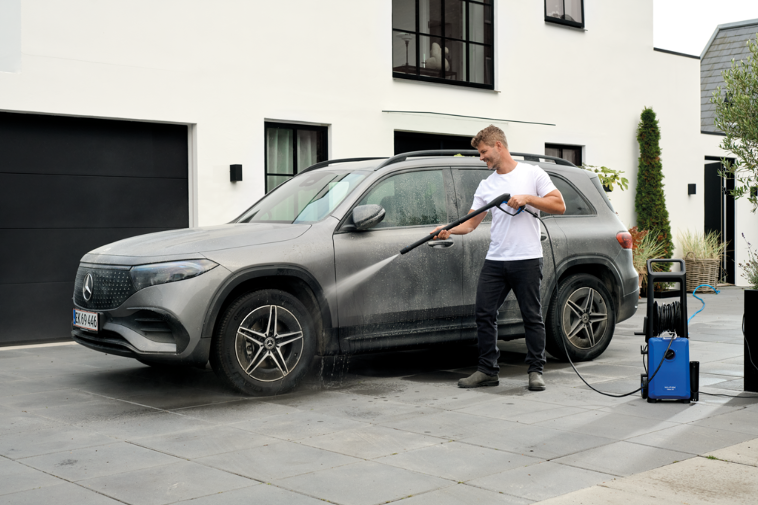 Man washes gray car with pressure washer in front of modern house.