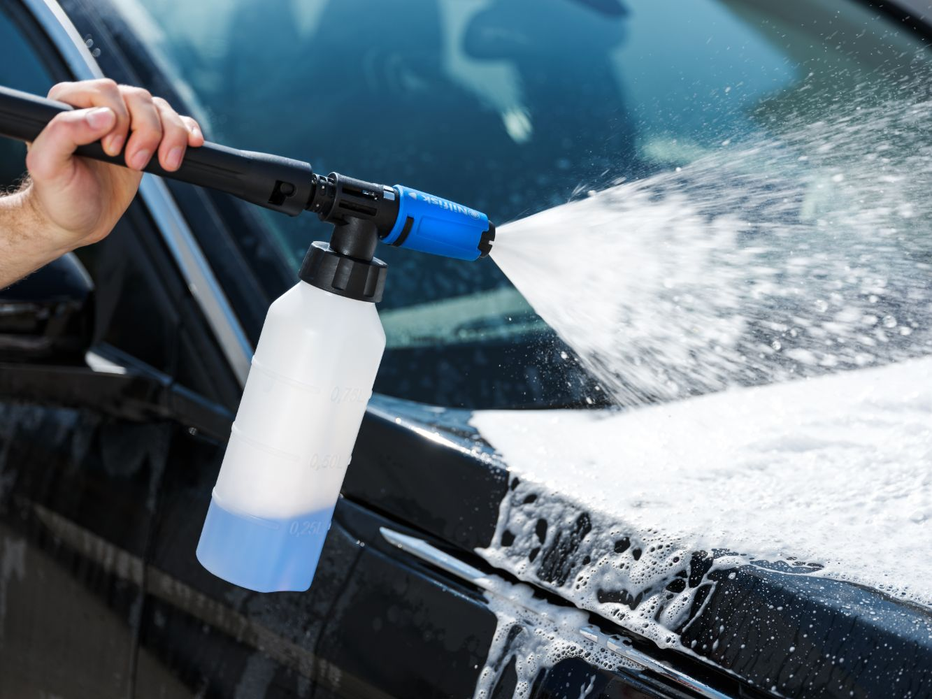 A hand holds a pressure washer with a foam bottle, spraying a car.