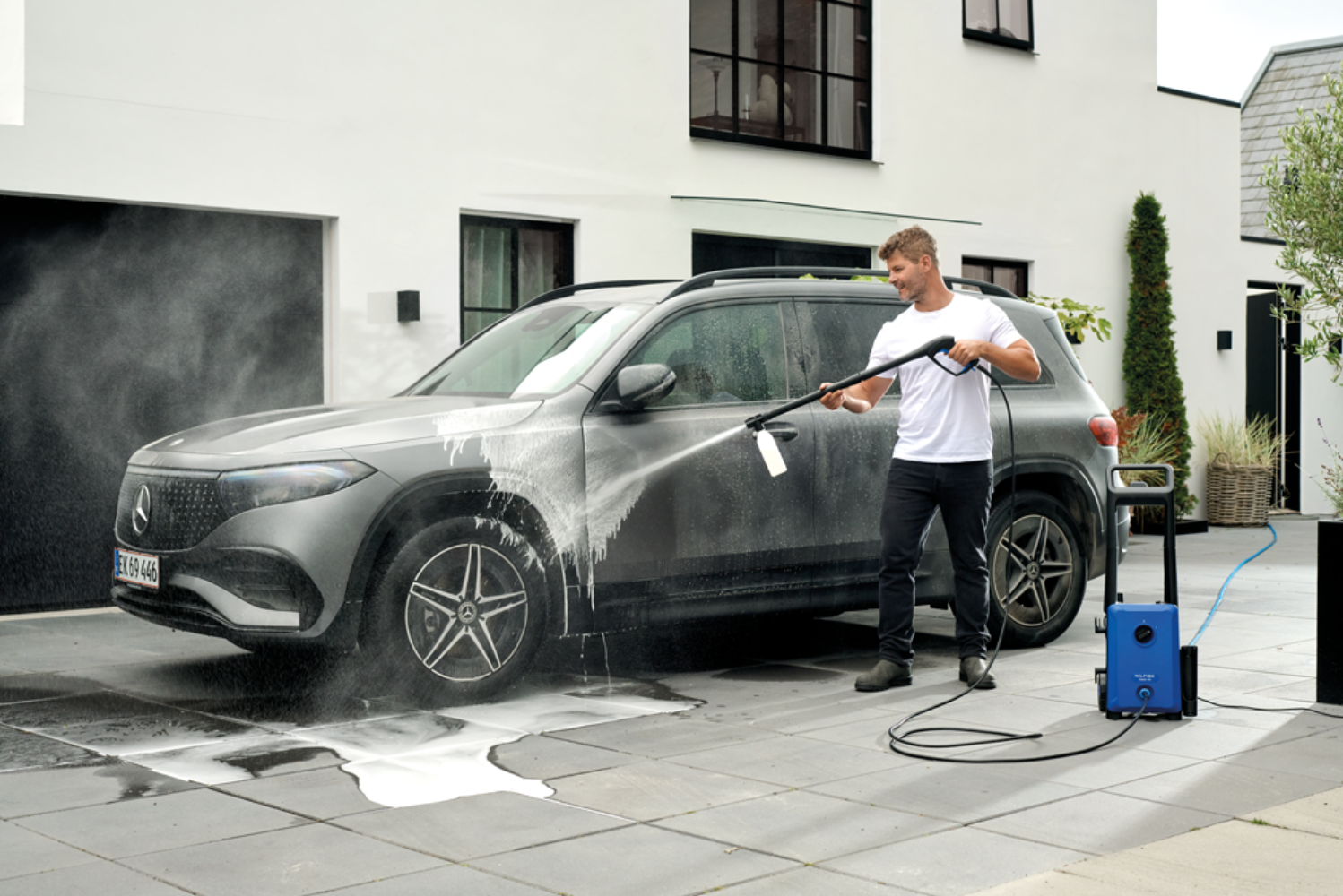 Man washing a gray car with a pressure washer on a paved driveway.
