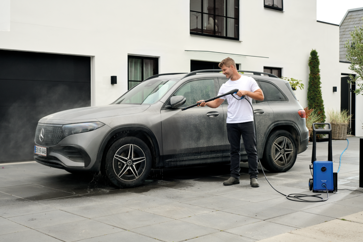 Man washes grey car with pressure washer in front of modern house.