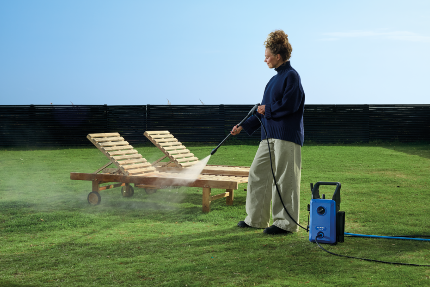 Woman pressure washing lounge chairs in a grassy yard.