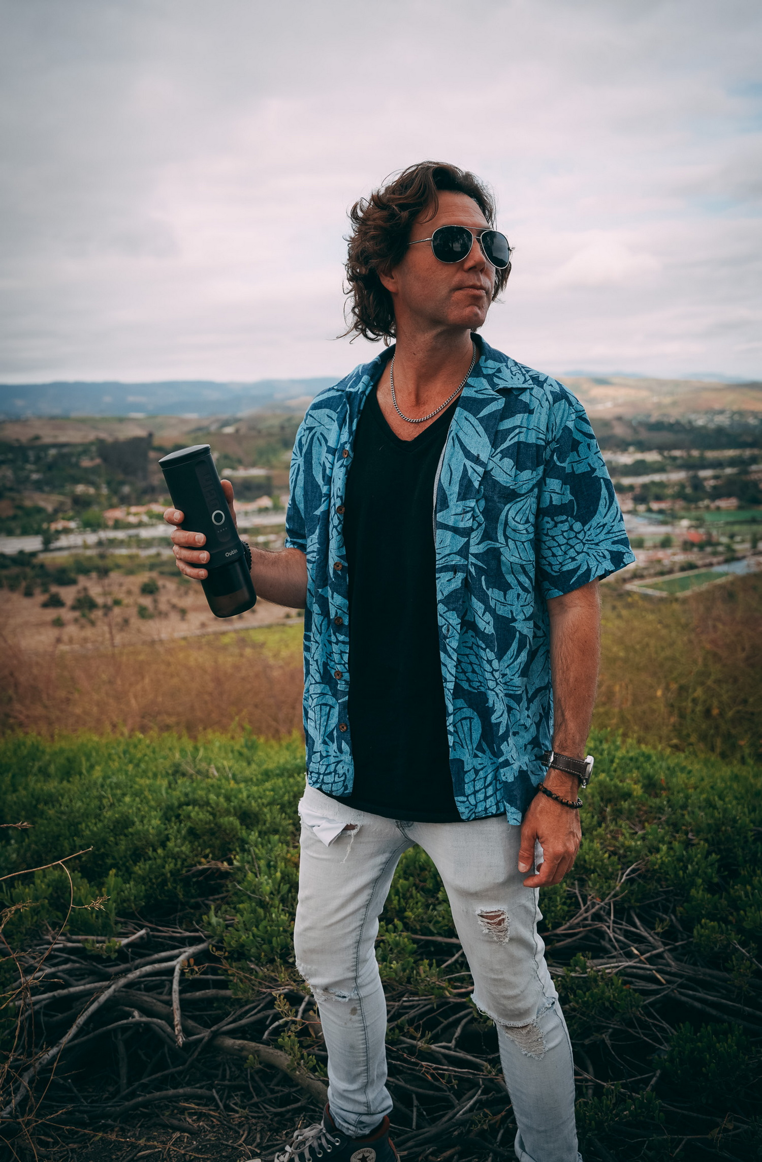 Mężczyzna z kręconymi włosami, okularami przeciwsłonecznymi, niebieską koszulą i podartymi dżinsami trzyma czarny walec.