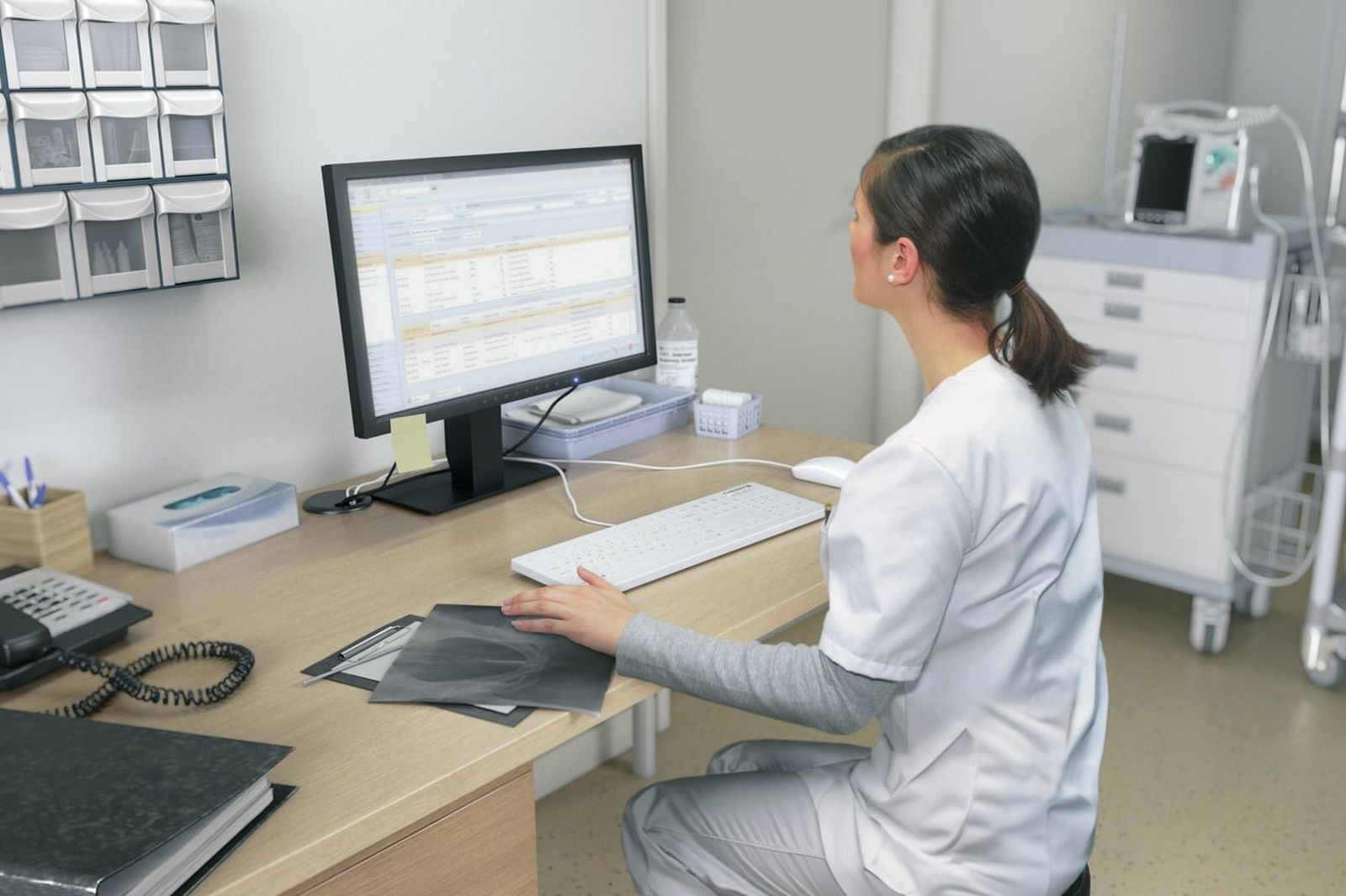 Eine Frau in weißer Arbeitskleidung sitzt an einem Schreibtisch und blickt auf einen Computerbildschirm.