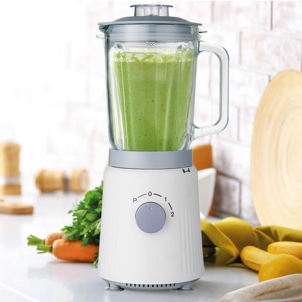 Biały blender z zielonym smoothie, obok marchewki i pietruszki.