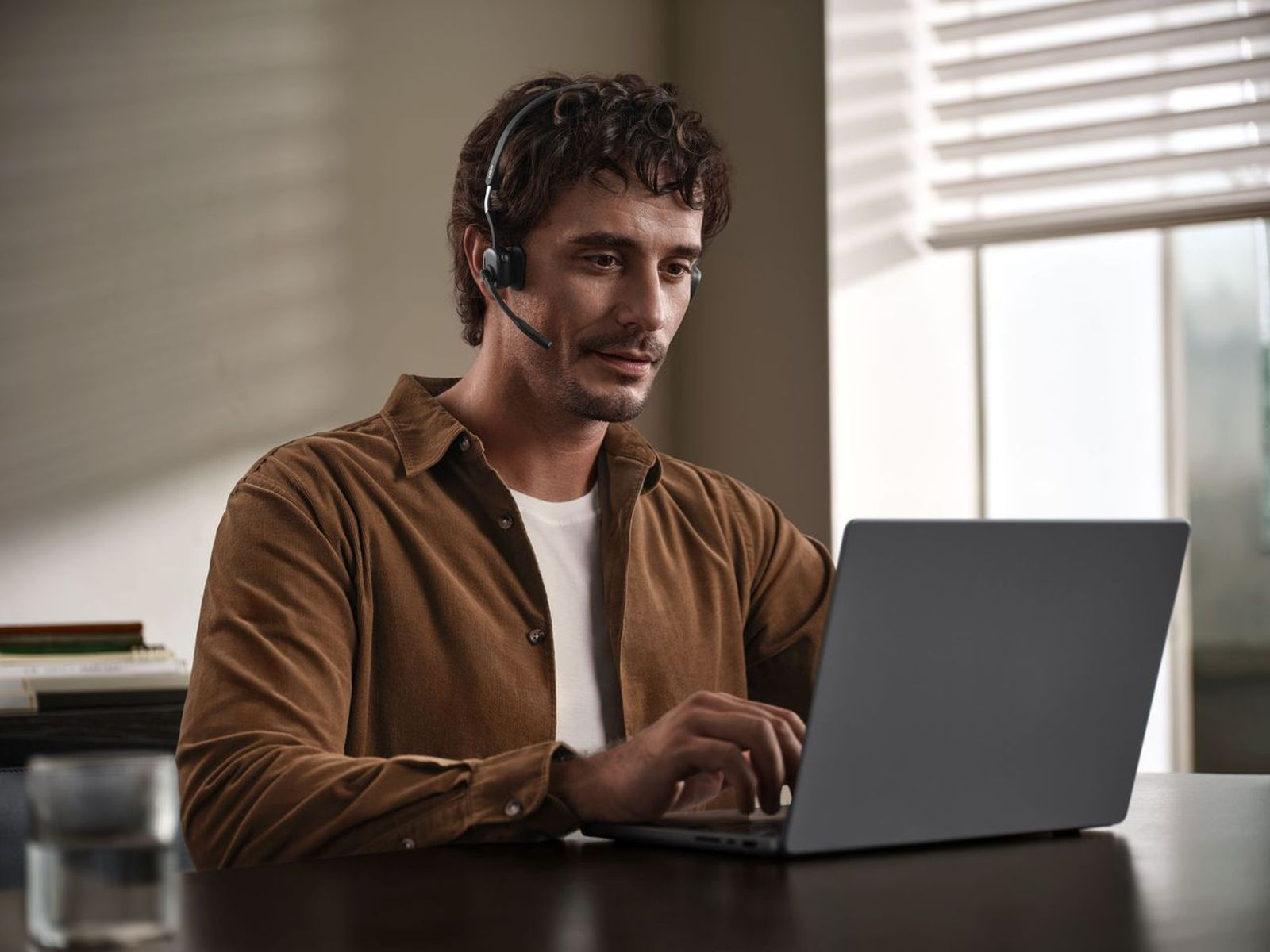 Mężczyzna w zestawie słuchawkowym pisze na laptopie. Nosi brązową koszulę.