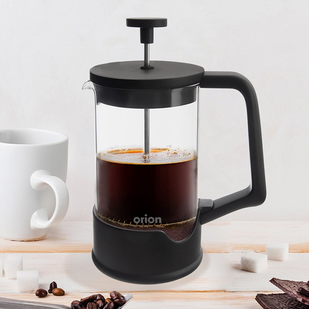 Una prensa francesa negra con café junto a una taza blanca sobre una mesa de madera.