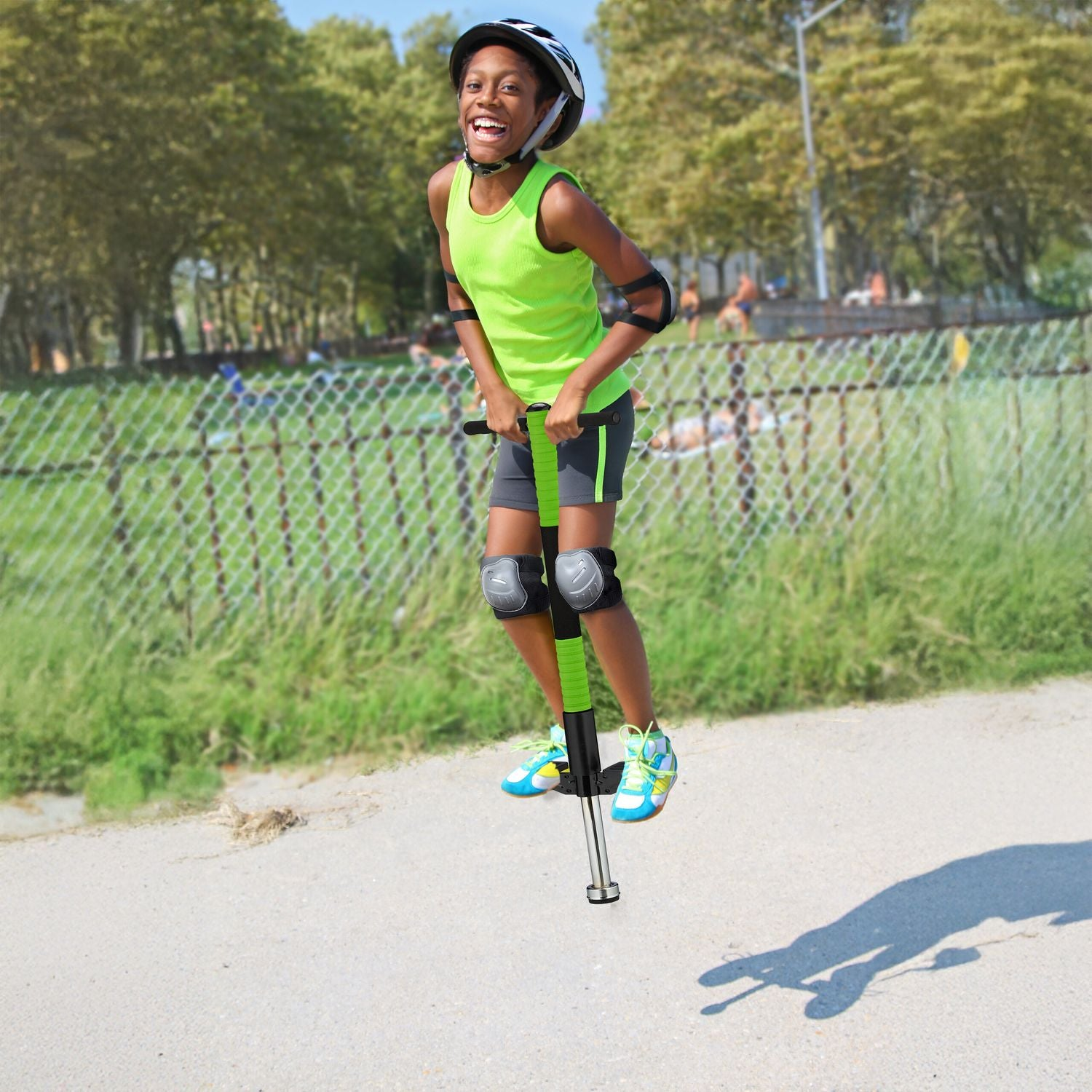 Un niño sonriente con casco, camiseta verde brillante y protectores salta en un pogo stick en un camino pavimentado.